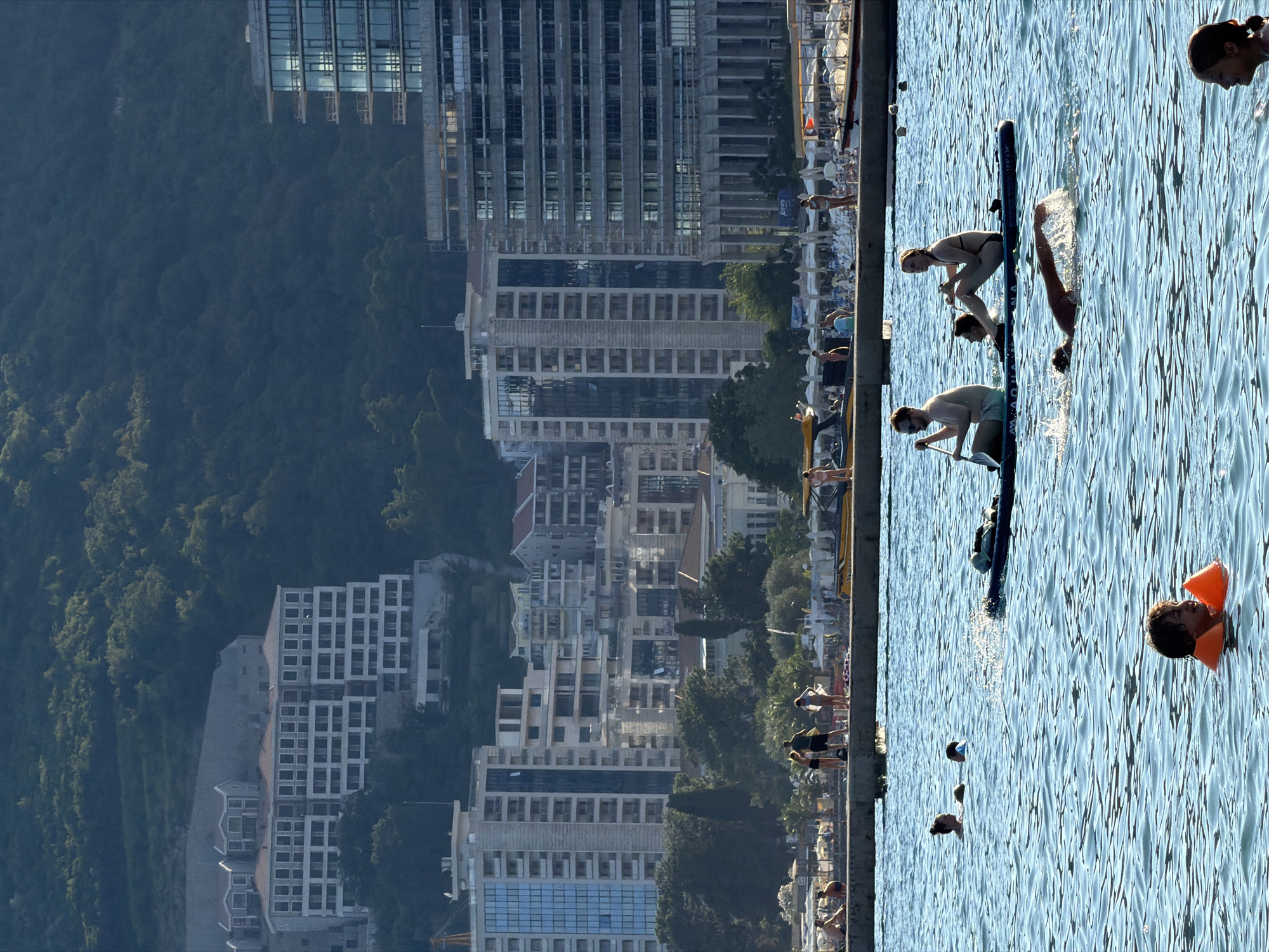 The image depicts a sunny day at a coastal area in Budva, Montenegro, with a mix of modern architecture and recreational activities. In the foreground, several people are enjoying the water. Two individuals are on a paddleboard, one paddling and the other seated, with another person swimming in front of them. A child wearing an orange life vest is also visible in the water, smiling.
In the midground, a concrete promenade separates the water from a beach area with numerous umbrellas and loungers. People are seen walking and relaxing on the promenade and beach. Beyond the beach, a dense line of trees forms a backdrop, behind which a series of multi-story buildings rise up the hillside. These buildings exhibit a variety of architectural styles, ranging from older, multi-gabled structures to more modern high-rises with extensive glass facades. The overall impression is one of a developed coastal resort with a bustling atmosphere.