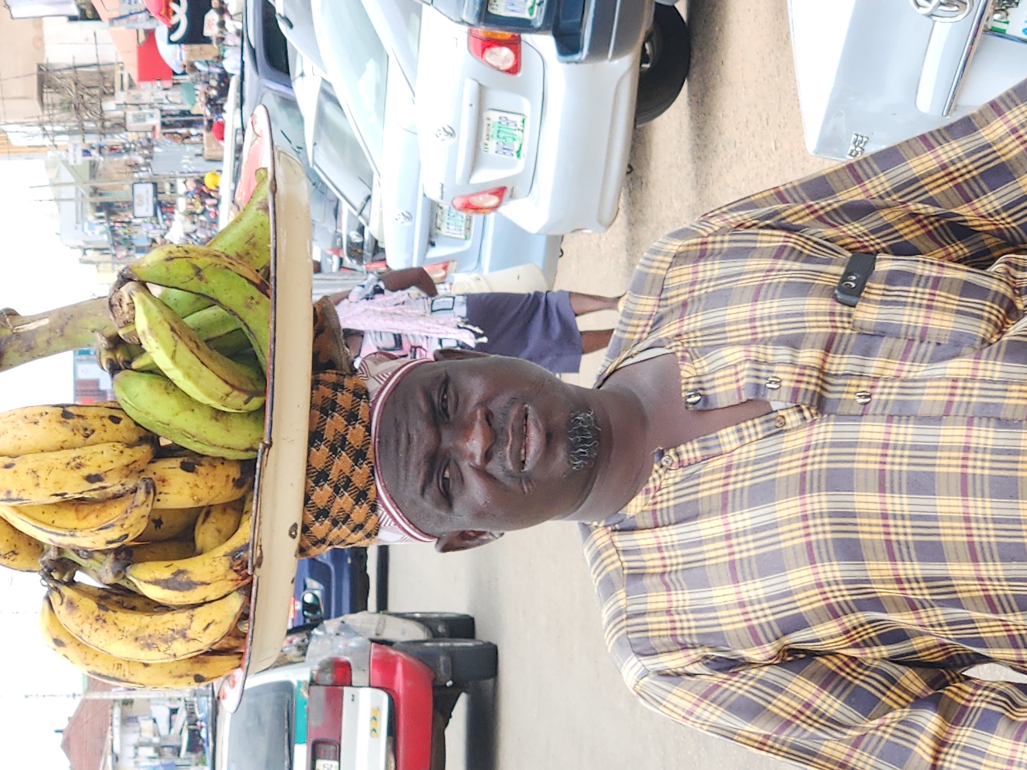 In the city of Jos, Nigeria, a man wearing a checkered shirt and a cap stands facing the camera. He has a goatee and a slightly worried expression. On his head, he carries a large tray filled with bananas. The tray is supported by a patterned cloth wrapped around his head and shoulders. Behind him, several cars are parked, suggesting a busy street or market area. In the background, buildings and people can be seen, indicating an urban environment. The overall scene depicts a vendor selling fruit in a bustling Nigerian city.