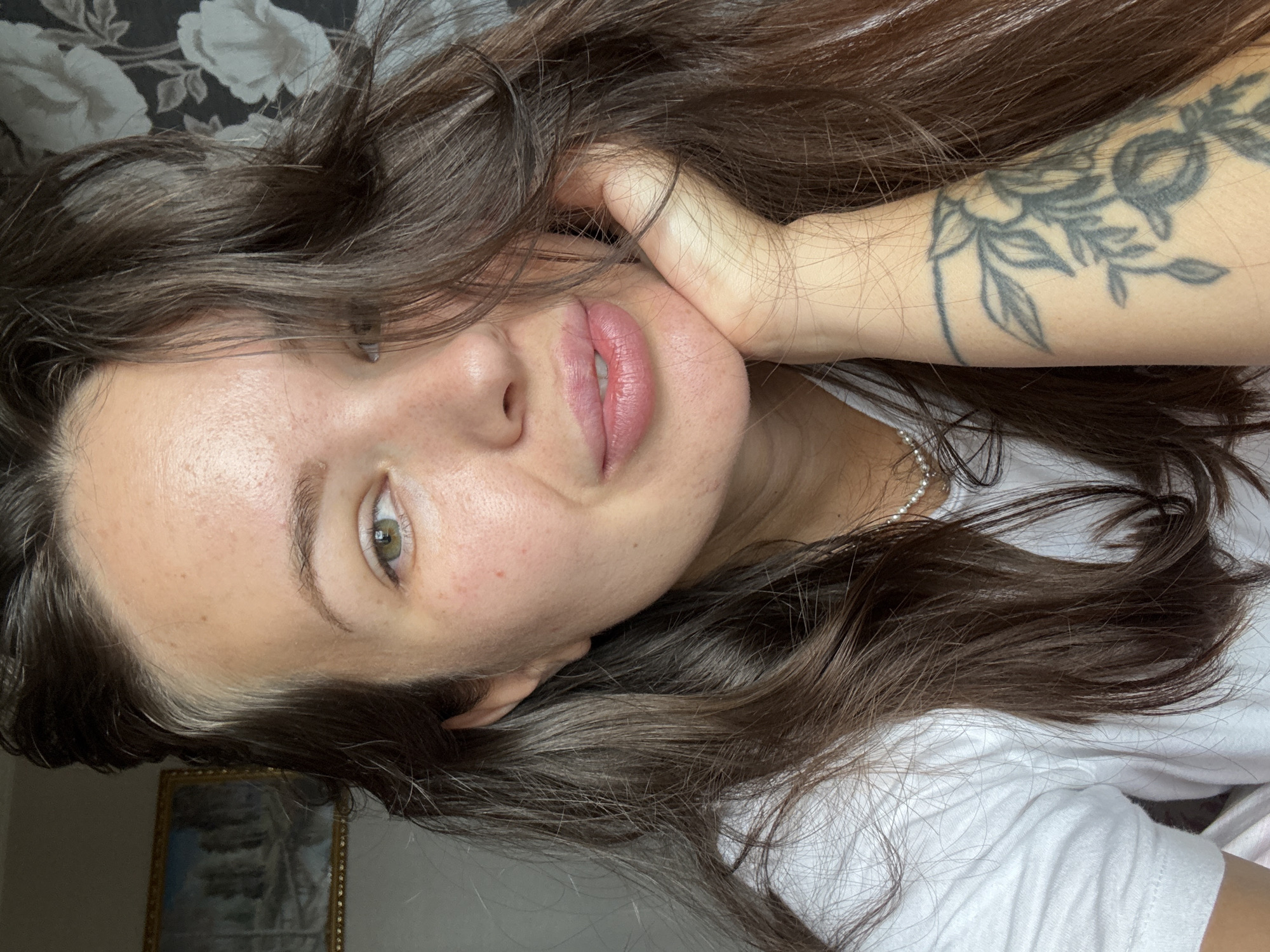 The image is a close-up, self-taken portrait of a young woman with long, dark brown hair. She is resting her chin on her right hand, which is near her mouth. Her hair is partially covering her face. She is wearing a white t-shirt and a pearl necklace. A tattoo with a floral design is visible on her left forearm. The background appears to be indoors, with a patterned wallpaper and a framed picture on the wall. There is no discernible indication of the image being taken in Bely Yar, Russia.