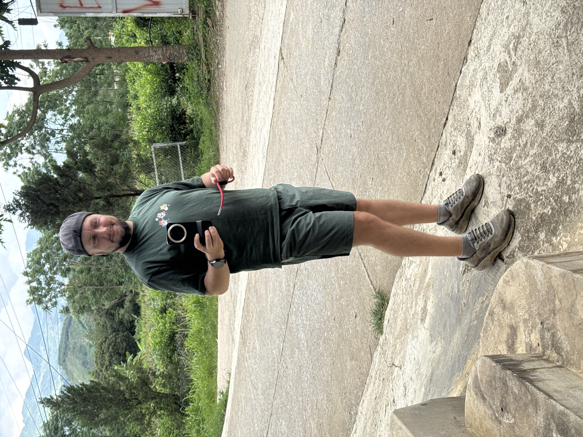 A man stands on stone steps in Trung Tam Ward, Vietnam. He is wearing a dark green t-shirt with cartoon images, dark green shorts, a grey cap, and grey sneakers. He is holding a black smartphone and a pair of red glasses. Behind him, lush greenery and a view of the valley are visible. The foreground shows a concrete path and stone steps.