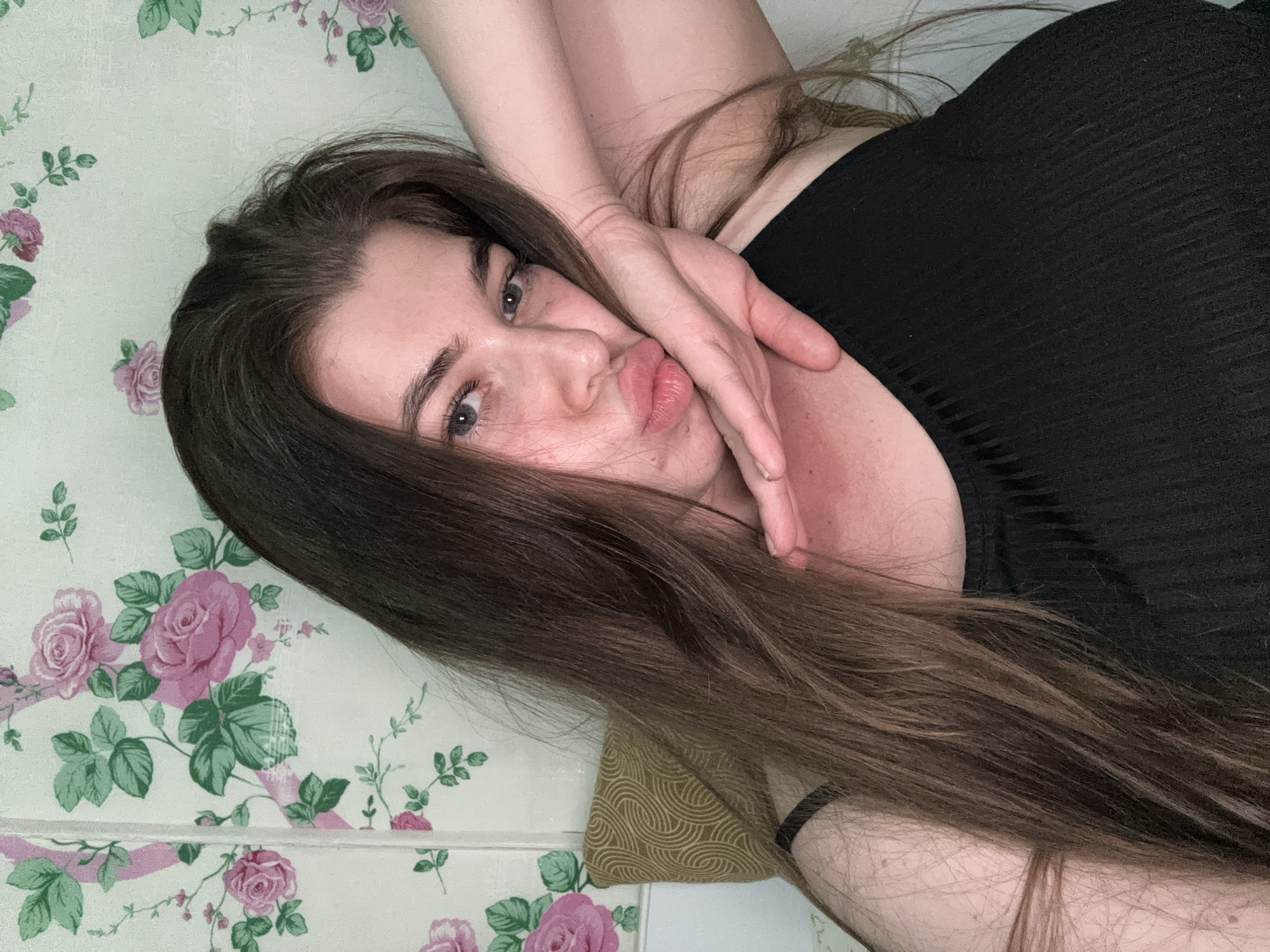 A young woman with long, brown hair is leaning into the camera, her hand resting on her cheek. She is wearing a black tank top. She has light-colored eyes and is making a kissing face. The background is a white wall with a floral pattern of pink roses and green leaves. A textured, patterned pillow is visible behind her left shoulder. The scene appears to be indoors, possibly a bedroom or living space. There is no indication of the location being in the city of Мещёры, Russia.