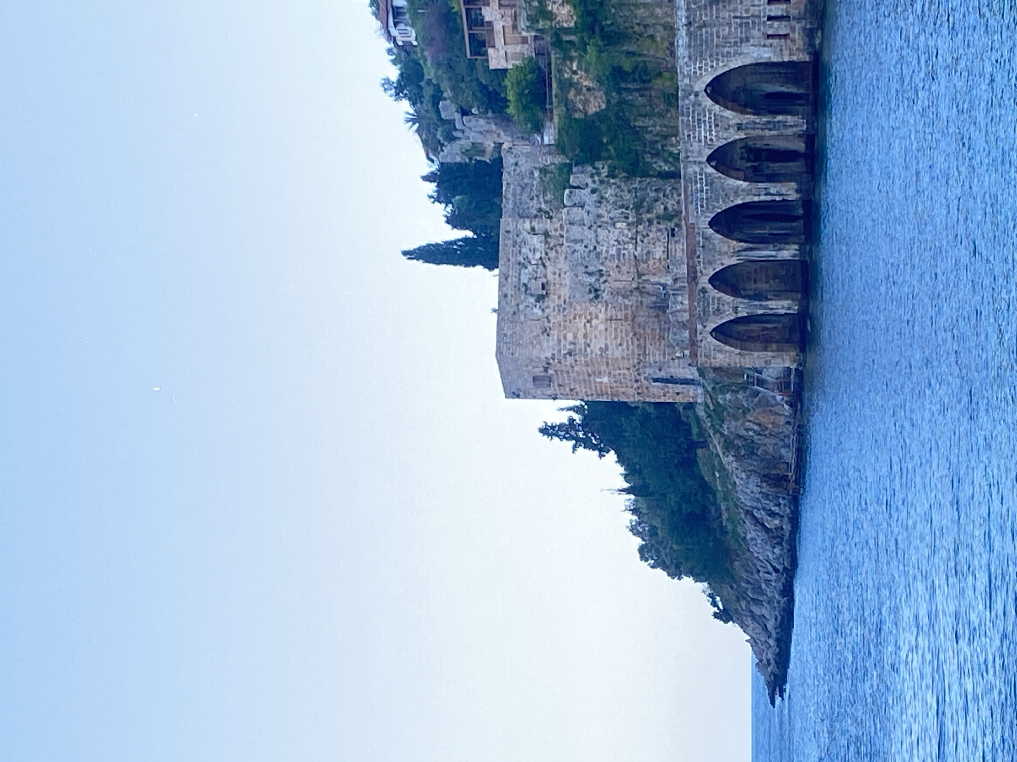 The image depicts a portion of the Alanya Castle walls in Alanya, Turkey, as viewed from the sea. The stone structure features multiple arched openings at its base, which appear to extend into the water. The wall rises vertically and supports vegetation on its upper levels. Cliffside vegetation is present along the rocky shoreline. To the left, the image captures a clear, light blue sky, while to the right, the sea extends with visible water ripples. The building structure has some residential houses built on the cliff above.