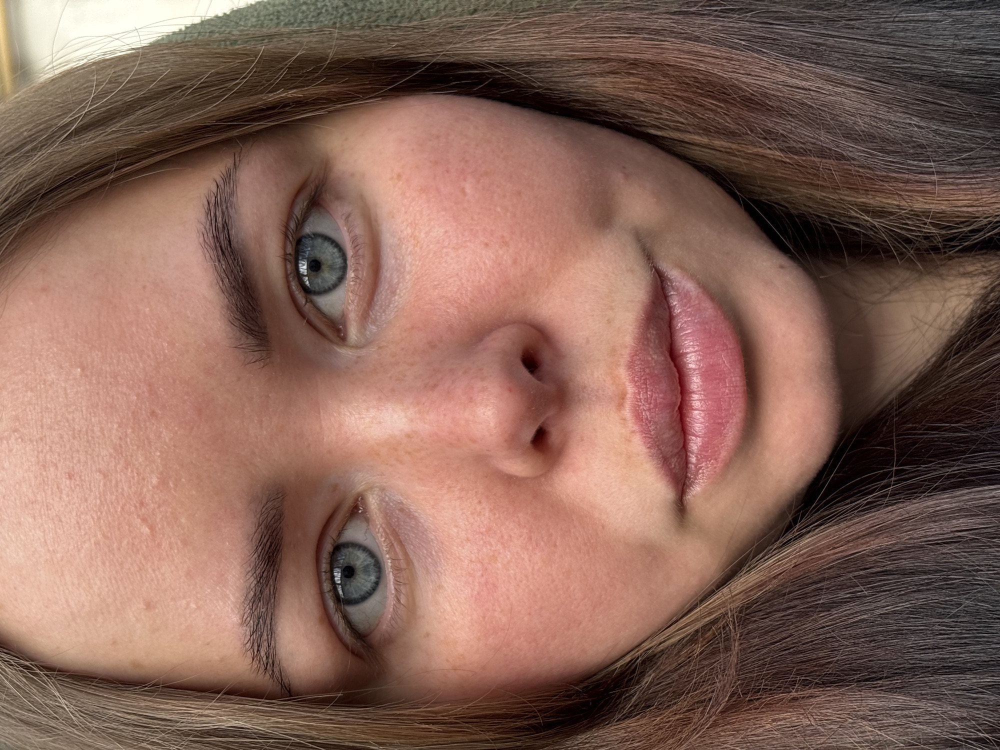 A close-up portrait of a young woman with fair skin and light brown hair is featured. Her eyes are a distinctive blue-gray color. She has freckles on her nose and cheeks. Her lips are pink and appear moisturized. No specific location in Izhevsk, Russia, is discernible from the image.