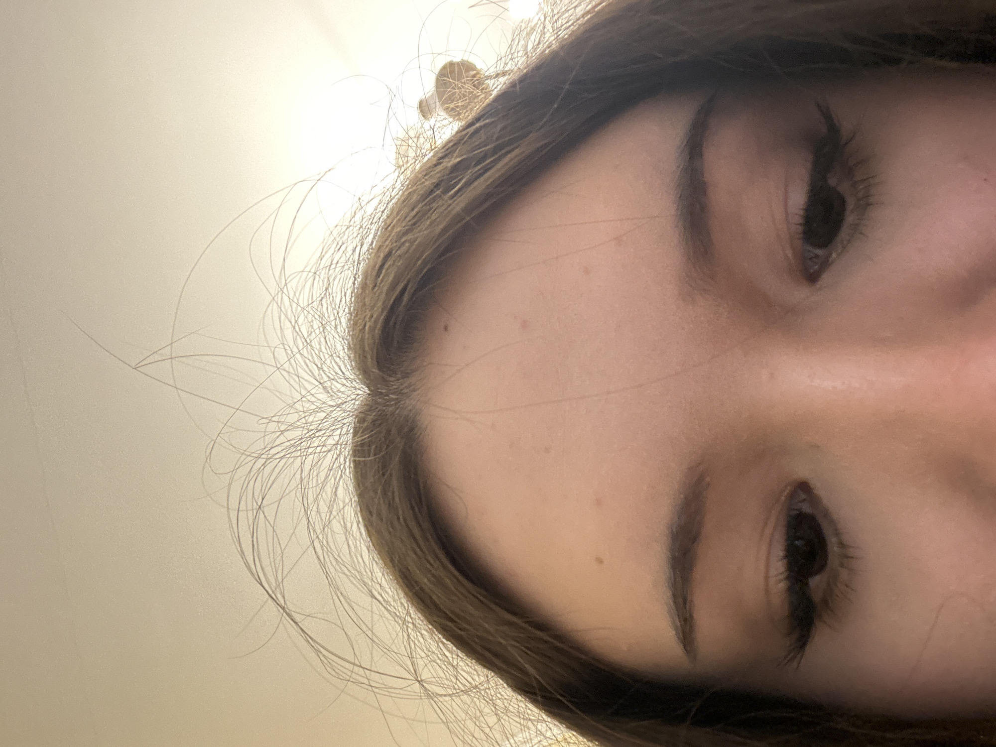 This close-up, low-angle image features the upper portion of a person's face, specifically the forehead, eyebrows, and eyes. The subject appears to be female, with dark brown hair framing the top of the forehead and around the temples. Wispy strands of hair are visible against the light background.
The perspective is unusual, looking upwards at the face, which is tilted back. Both eyes are visible and appear dark, with long, dark eyelashes. The eyebrows are dark and well-defined. The skin on the forehead shows some natural texture and a few small moles.
The background is a plain, light-colored surface, likely a ceiling or wall, illuminated by a bright light source visible in the upper right portion of the frame, casting a soft glow. A small, light-colored, textured object or decorative element is partially obscured by hair in the upper left, possibly part of a light fixture or room decor.
The scene takes place indoors, likely under artificial lighting, in Naberezhnye Chelny, Russia. No specific activity is discernible beyond being photographed in a close-up portrait. Emotions are difficult to interpret due to the extreme angle and partial view of the face. There is no visible text in the image.