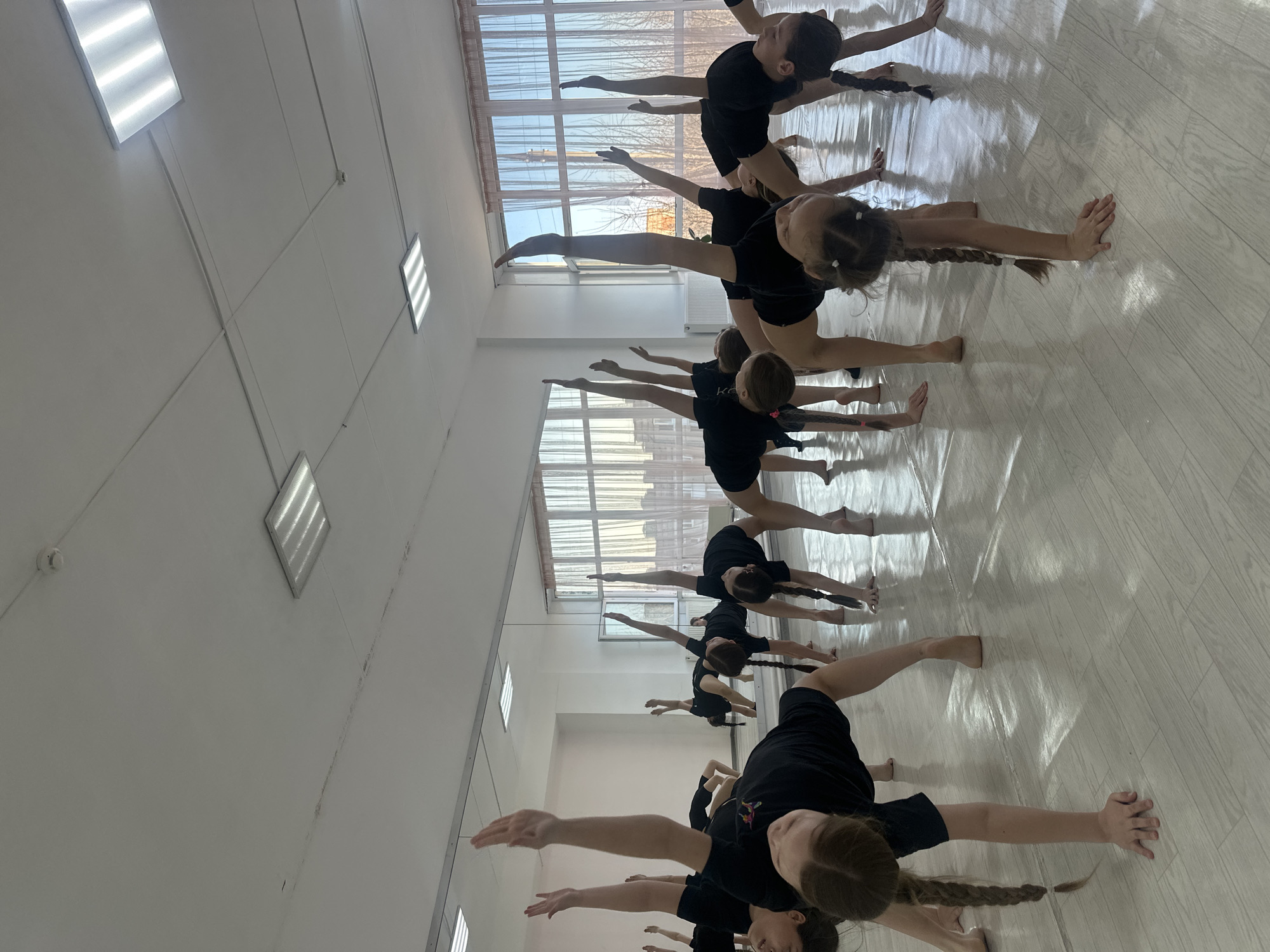 A dance class is being held in a studio in Votkinsk, Russia. The room is brightly lit with rectangular ceiling lights. A long row of young dancers wearing black outfits is arranged in a line, performing a stretching exercise. They are on their hands and feet, with legs split to the side. A window with light curtains is on the wall behind them. The floor is light-colored wood.