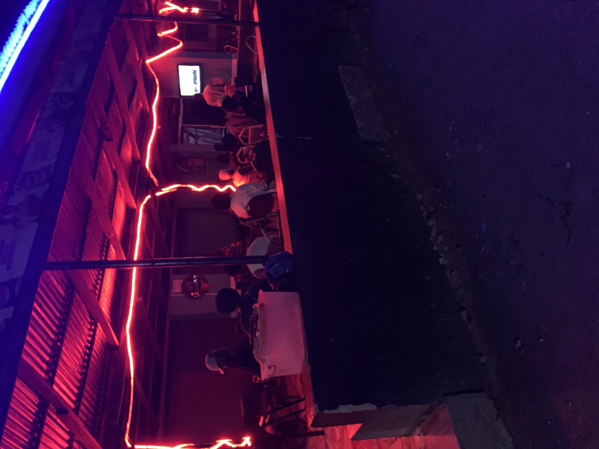 The image depicts an outdoor establishment in New Stadium, Nigeria, illuminated by red neon lights. A corrugated metal structure serves as a roof. A group of people are seated inside, watching a television. Outside, the surroundings are dark.