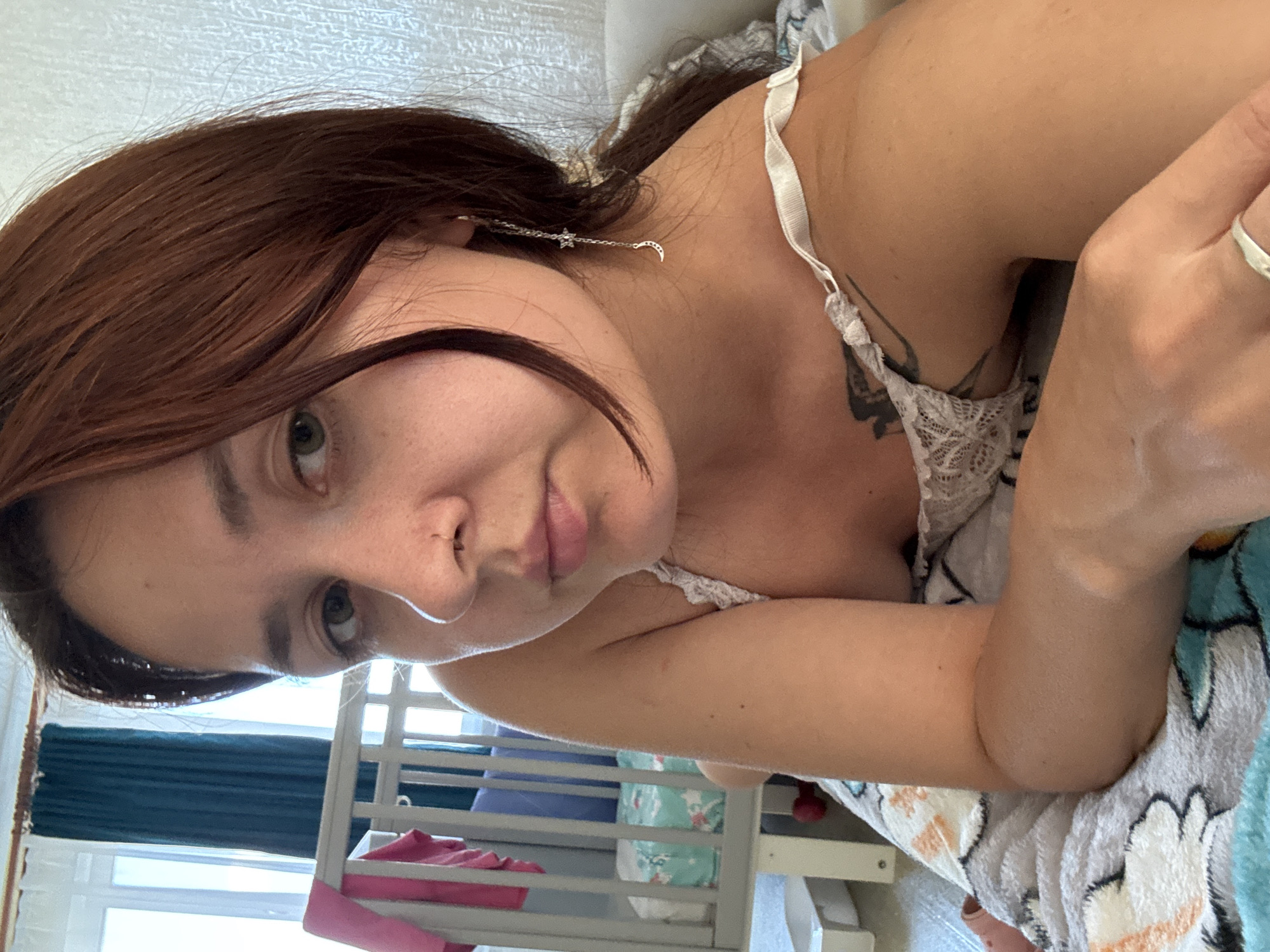 A young woman with auburn hair and green eyes is lying on a bed, looking towards the camera. She is wearing a white lace-trimmed top and has a silver moon and star earring in her left ear. A tattoo is visible on her left shoulder. The background includes a crib, teal curtains, and a window. The scene appears to be indoors, likely in a bedroom. There is no information in the image that would indicate the specific location in Naberezhnye Chelny, Russia.