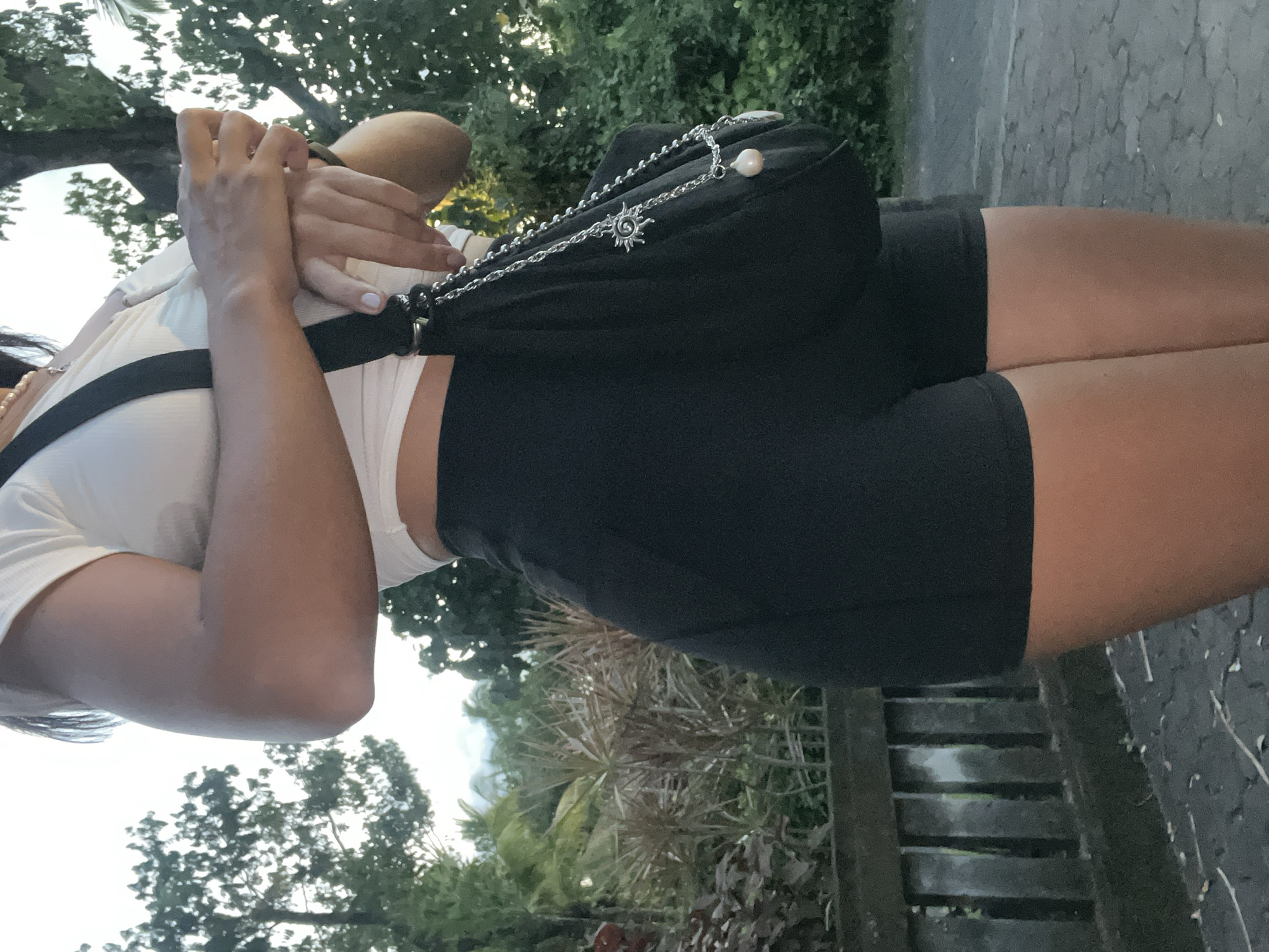 A vertically oriented, eye-level photograph shows an individual in Kuta Selatan, Indonesia, with a dark purse, fair skin, short black pants, and a white top, standing outdoors on a partially paved area near a green space. The individual is seen from the torso down, facing slightly to the right. The background features dense green vegetation and a portion of a gray stone or cement structure on the right. The lighting appears soft and diffused.
