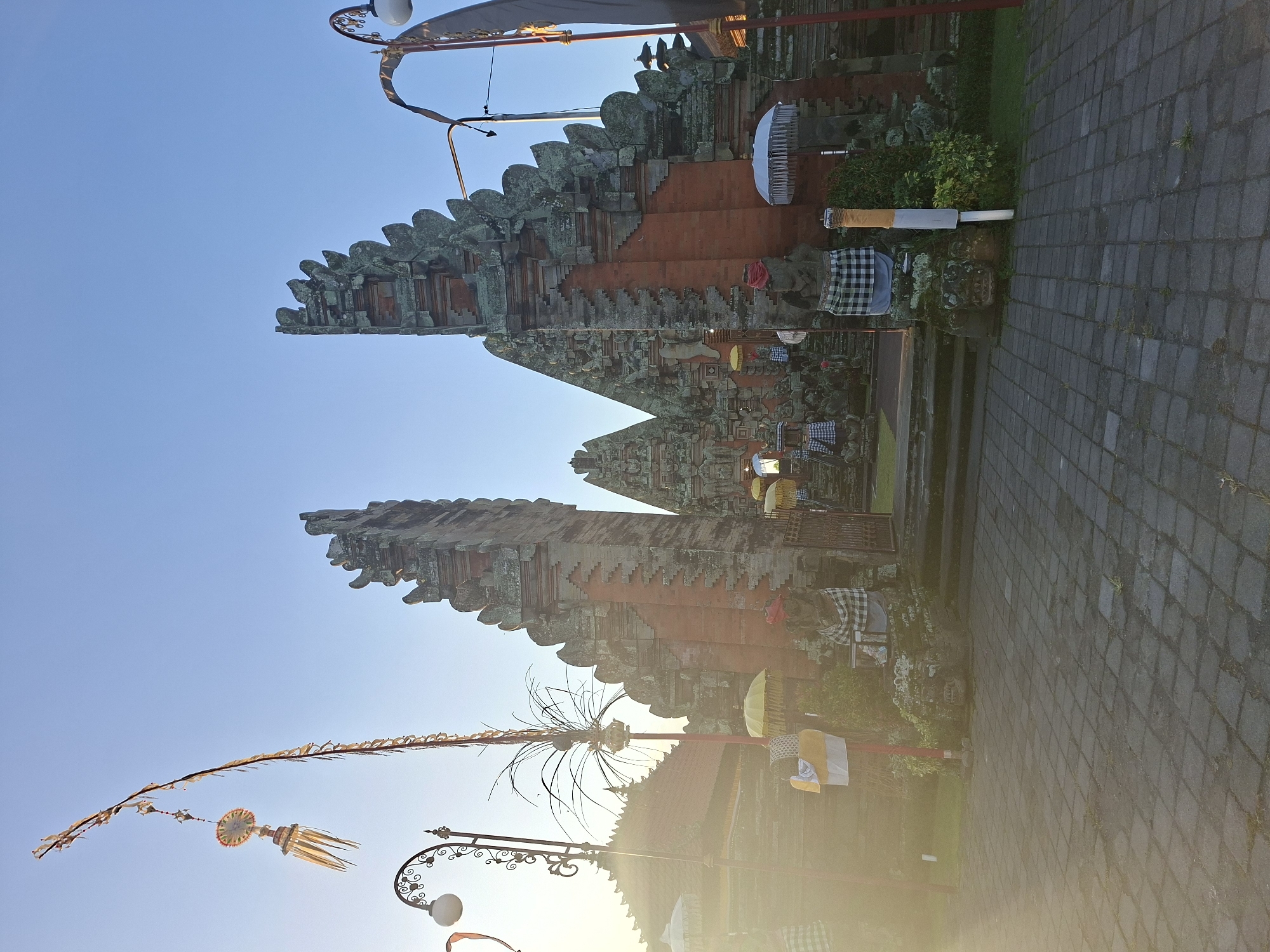 This image depicts the entrance to a traditional Balinese temple, likely in Sukawati, Indonesia, under a clear blue sky during daylight hours. The sun's bright light casts a lens flare effect across the left side of the frame, creating a warm glow and obscuring some details.
The central focus is a towering split gate, constructed from brick and stone, adorned with intricate carvings and moss. The gate's jagged, tiered roofline is characteristic of Balinese architecture. Flanking the entrance are two stone statues, dressed in traditional checkered cloths and red headbands, which appear to be temple guardians.
In front of the gate, a paved courtyard made of dark stone slabs leads towards the entrance. Several yellow and white umbrellas are strategically placed around the statues and the temple grounds, adding decorative elements. Tall, slender poles topped with decorative banners and ornaments are erected to the left and right of the main gate, further enhancing the festive or ceremonial atmosphere.
Vegetation, including green bushes and plants, is visible around the base of the temple structures and the statues, suggesting a well-maintained environment. A street lamp with a decorative curved arm and a white globe is positioned on the left side of the image, hinting at the presence of nearby amenities or infrastructure. The overall scene conveys a sense of cultural heritage and tranquility, characteristic of religious sites in Bali.