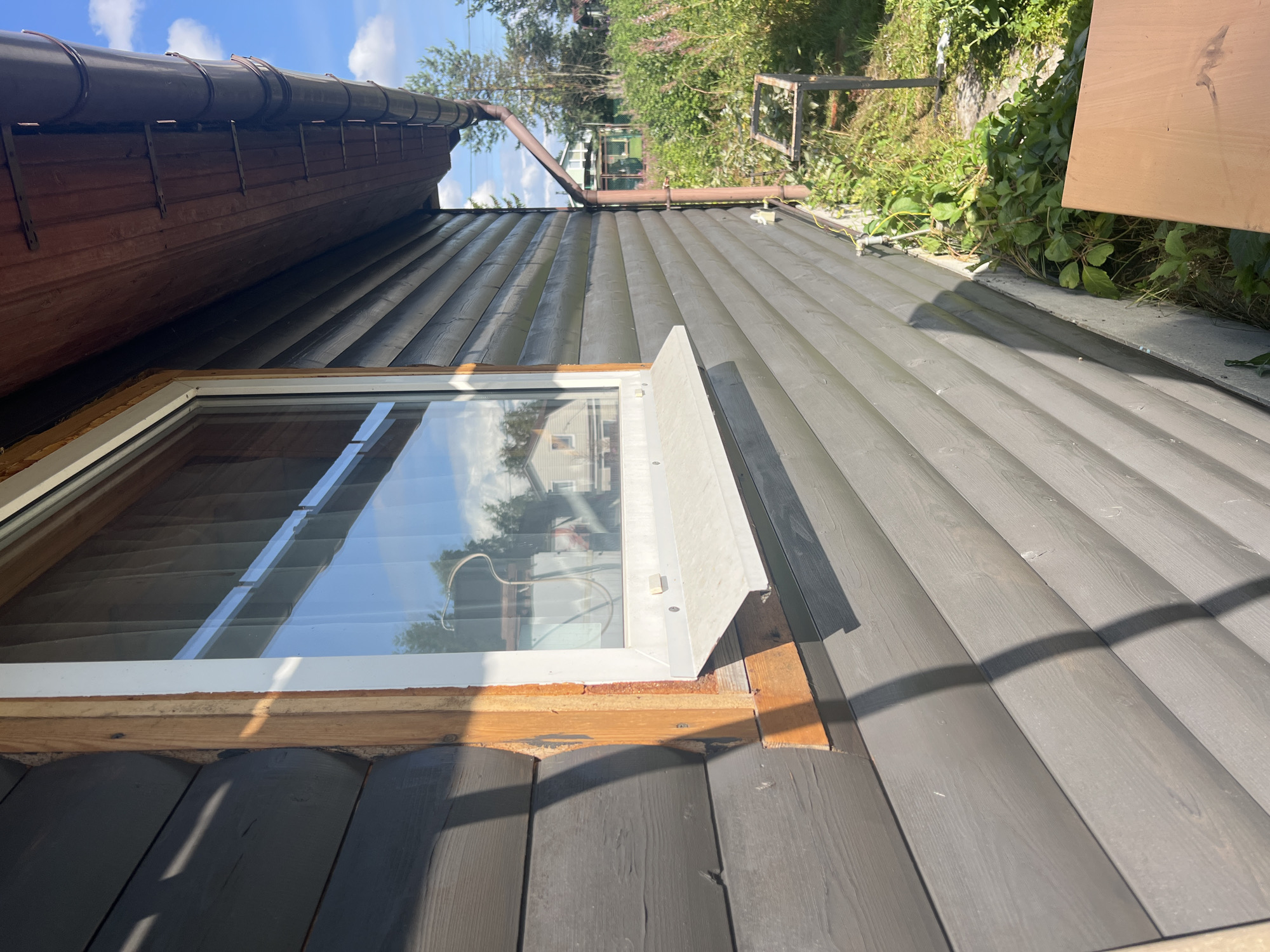 The image shows a close-up exterior view of a dark gray wooden log house with a white-framed window. The window has a white sill and a reflection of the sky and trees is visible in the glass. The wooden logs are horizontal and have visible grain. Part of a brown gutter system is visible at the top of the house. To the right of the house, there is overgrown green vegetation, including tall flowering plants, and a metal frame structure on the ground. A brown downspout extends down the side of the house. The scene appears to be in a rural or suburban area, likely a dacha or country house, given the vegetation and other houses visible in the background. The lighting suggests it is daytime, with sunlight casting shadows. The location is identified as Роса Снт, Russia.