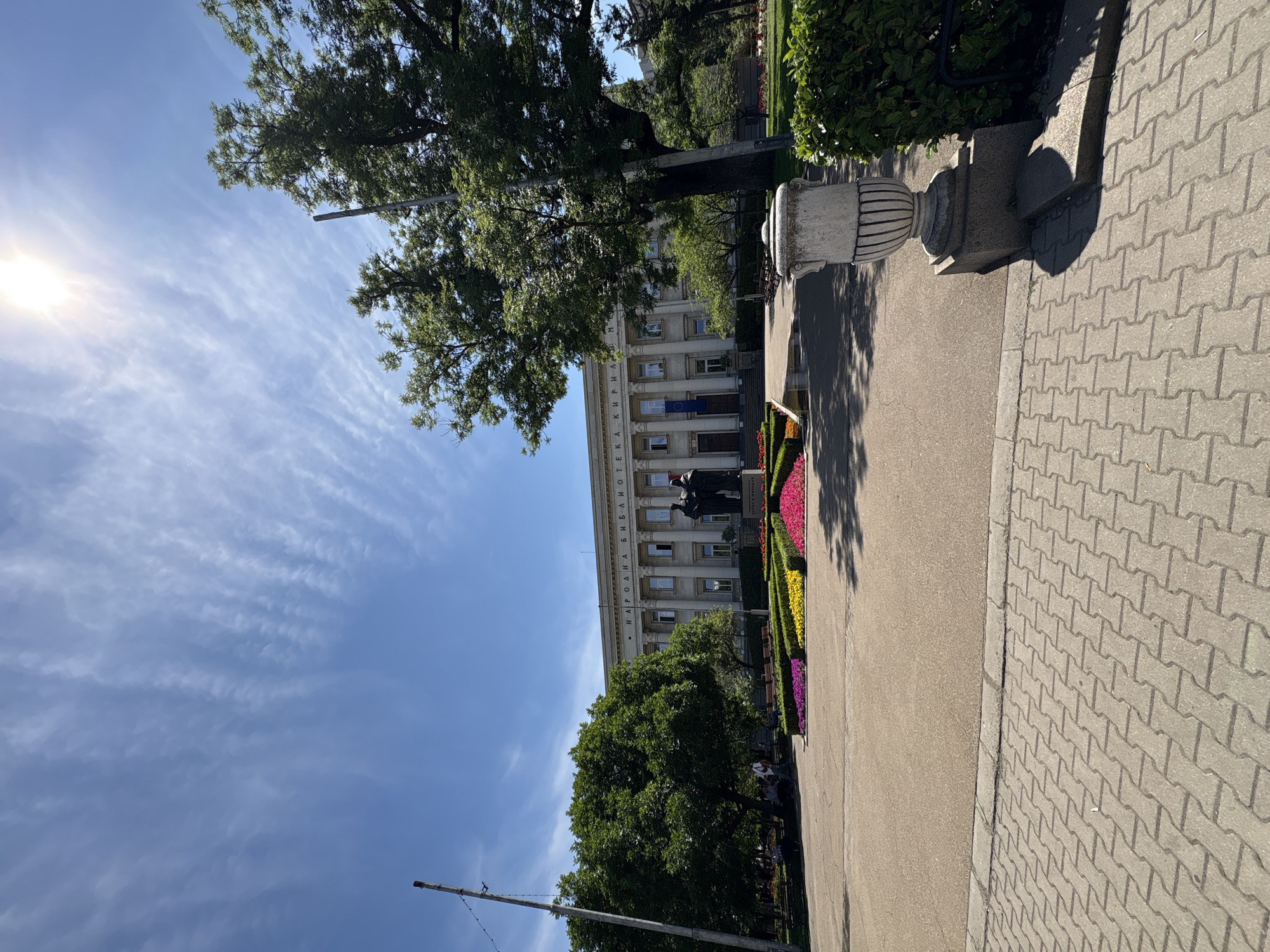 The image shows the National Library "Ivan Vazov" in Sofia, Bulgaria, on a sunny day. The building is large and classical in style, with columns and a pediment. In front of the library, there is a large flowerbed with colorful flowers. There are also trees and a statue of two figures. The foreground shows a paved walkway and a park area. The sky is blue with some wispy clouds. The sun is shining brightly, casting shadows. There are a few people in the park, some sitting on benches and others walking.