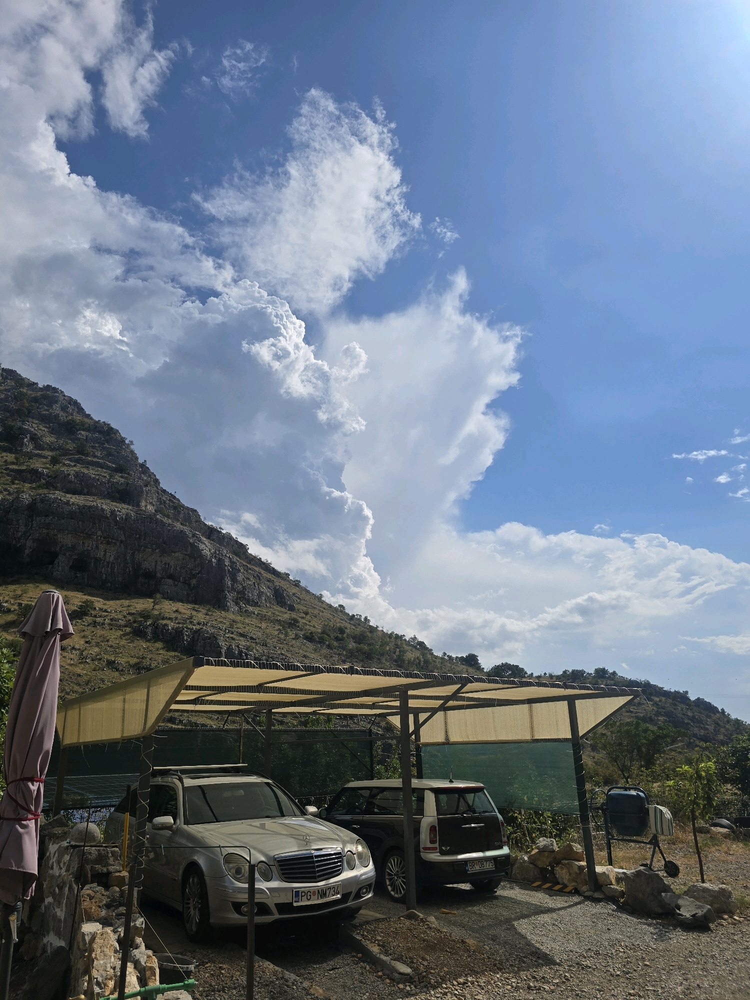 This is a medium-shot, eye-level, outdoor, vertical photograph taken on a sunny day with dramatic clouds. The scene depicts a parking area with two vehicles under a carport. In the foreground, a silver Mercedes-Benz E-Class sedan is parked facing forward, with its license plate visible as "PG NM 734". To its right, a dark-colored Mini Countryman hatchback is parked, facing away from the viewer, with its license plate reading "BR 08775". The carport has a tan fabric roof supported by metal poles. To the left of the carport, a folded light pink umbrella is leaning against a stone wall. Behind the carport, a green mesh fence is visible, and beyond that, a grassy slope dotted with trees leads up to a rocky mountainside. The sky is a vibrant blue with large, billowing white clouds, particularly prominent in the upper left and right quadrants. The overall impression is one of a rural or suburban setting, likely on the outskirts of Podgorica, Montenegro, given the license plate format.