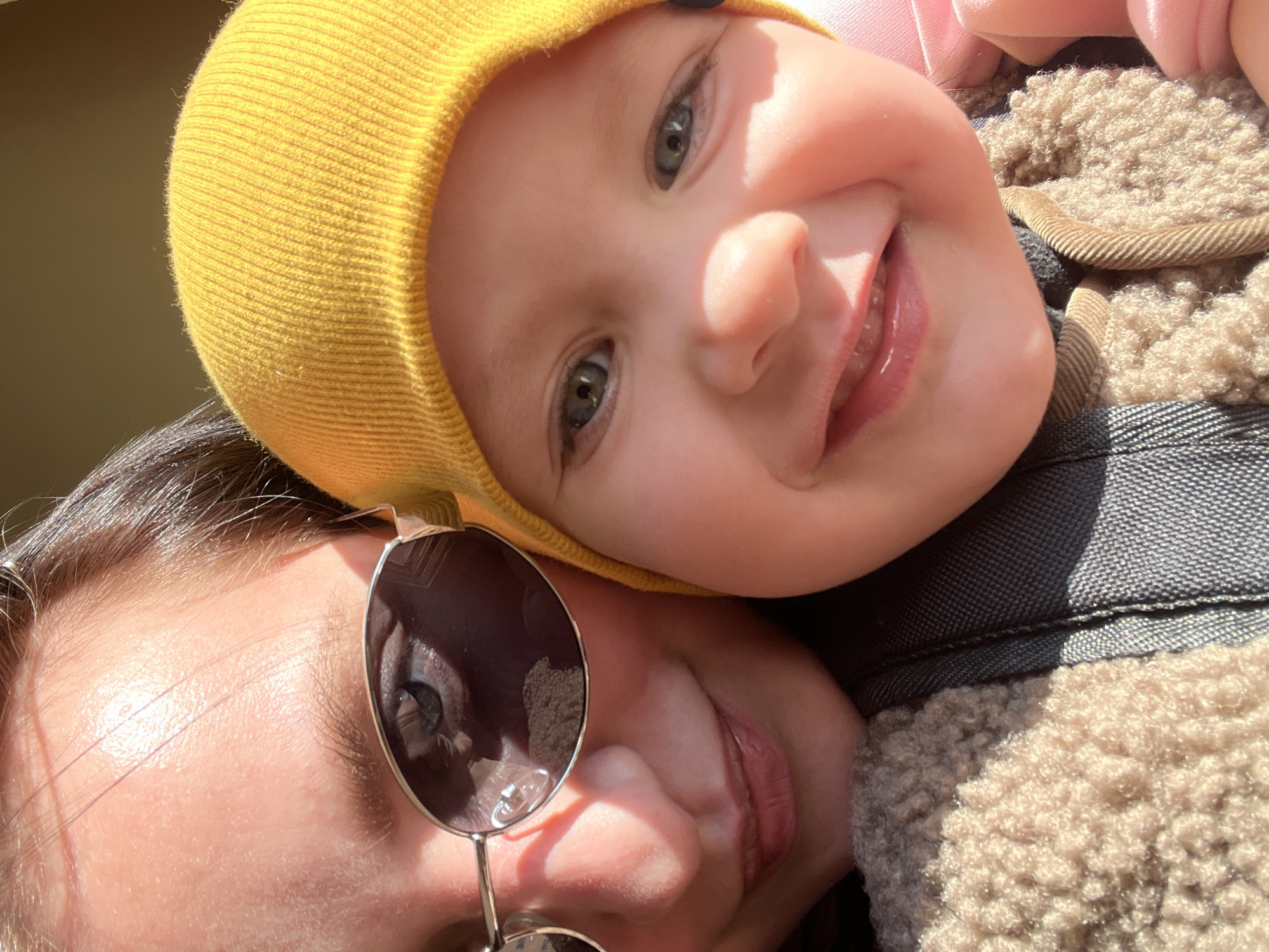 The image shows a close-up of a woman wearing sunglasses holding a baby. The baby is wearing a yellow beanie. The woman is wearing a jacket with a shearling collar. The background is out of focus. There is no discernible information from the image that identifies the location in the city of Марковский, Russia.