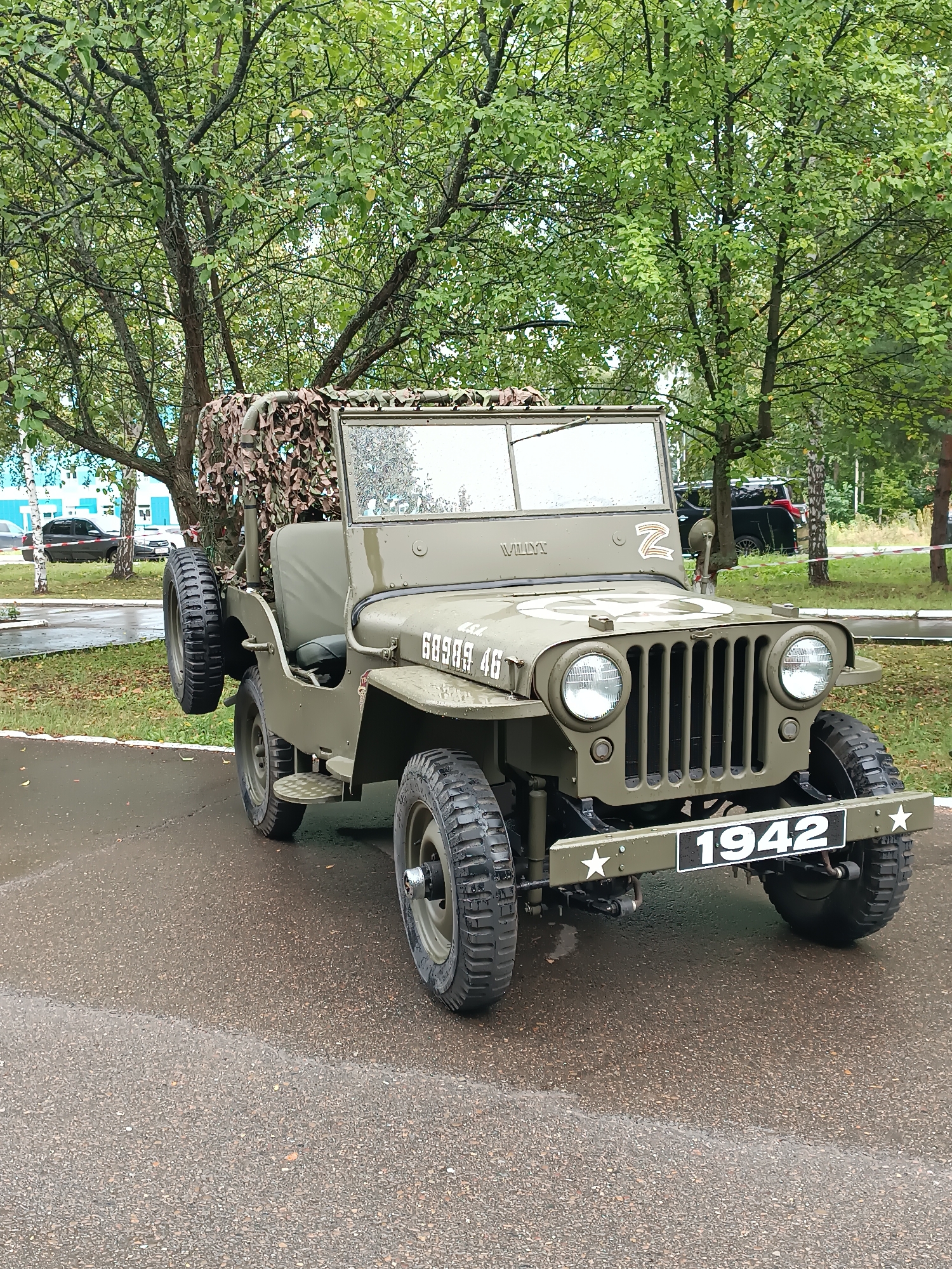 A parked, olive-drab military jeep, identified by the number "1942" on its license plate and a white star emblem, is the primary subject of this outdoor photograph. The jeep is positioned on a wet asphalt surface, with trees and foliage forming the background. The sky appears overcast, contributing to the wet appearance of the ground. In the background, a building with light-colored walls and windows is visible, along with a few parked cars, one of which is black. The presence of the number "Z" on the jeep's hood and a white star emblem suggests it might be a prop or a replica, possibly related to a patriotic event or display. The image appears to have been taken in Chaikovsky, Russia, given the context provided. There are no people visible in the frame.