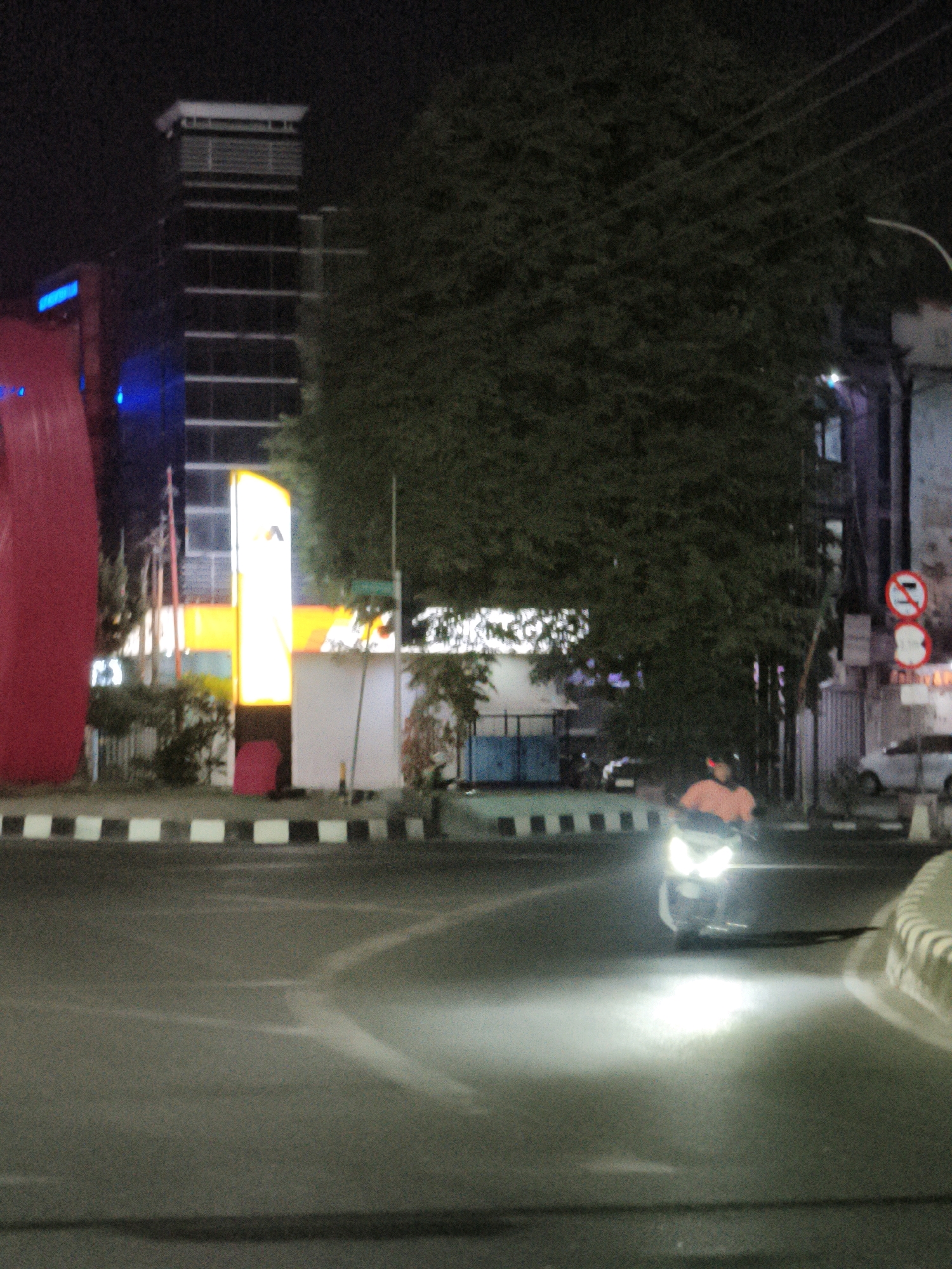 The image captures a street scene at night in Samarinda, Indonesia, likely near the Samarinda Central Plaza or a similar commercial area given the presence of a modern building and bright signage. A tall, dark building with horizontal window lines stands prominently in the background, illuminated by internal lights and possibly external lighting. To its left, a large, curved, red structure is partially visible, suggesting a large art installation or architectural feature. In front of the tall building is a bright, vertically oriented, illuminated sign with yellow and white elements, displaying a logo that appears to be a stylized 'A'.
The street itself is paved, with a distinct black and white checkered curb line running along the side. In the foreground, a motorcycle is being ridden by an individual wearing an orange shirt. The motorcycle's headlight is on, casting a bright beam of light across the asphalt and illuminating the road surface. The rider is captured mid-motion, turning on the street. In the background, other elements of the urban environment are visible, including trees, other buildings, and street signs. One sign, partially obscured by foliage, appears to be a red circular sign with a white horizontal bar and a diagonal line through it, commonly indicating a prohibition. Another white sign with a red border and a symbol of a cigarette with a diagonal line through it is also present, indicating "no smoking." A white car is parked on the right side of the road in the background. The overall atmosphere is dark, typical of an urban night, with artificial lights providing the primary illumination.