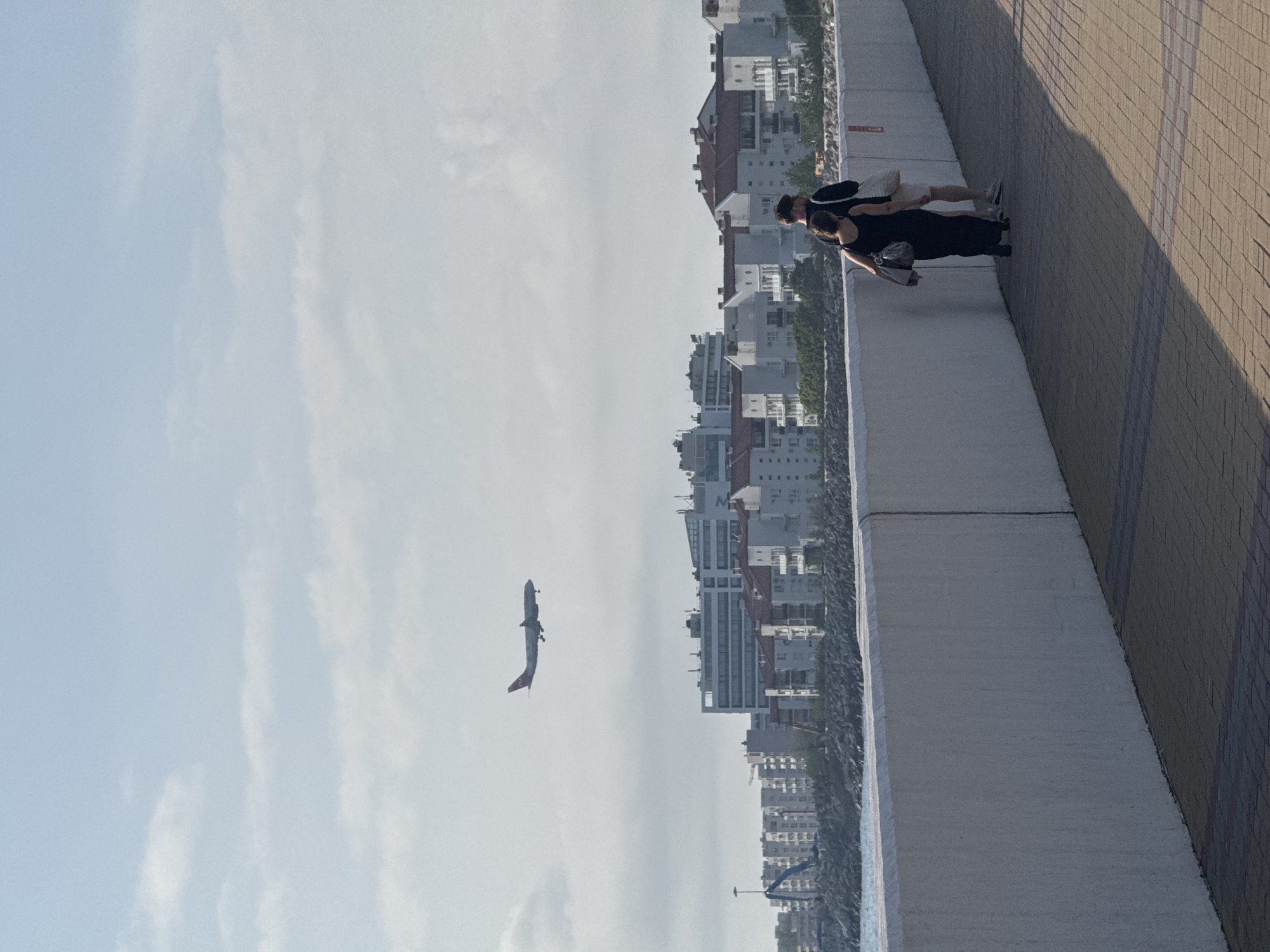 The image shows a scene from the city of Sirius, Russia. The sky is light blue with some clouds, and an airplane is in flight. On a raised sidewalk, two people are looking at something in the distance. The sidewalk is made of light-colored bricks. A series of buildings stretch along the horizon.