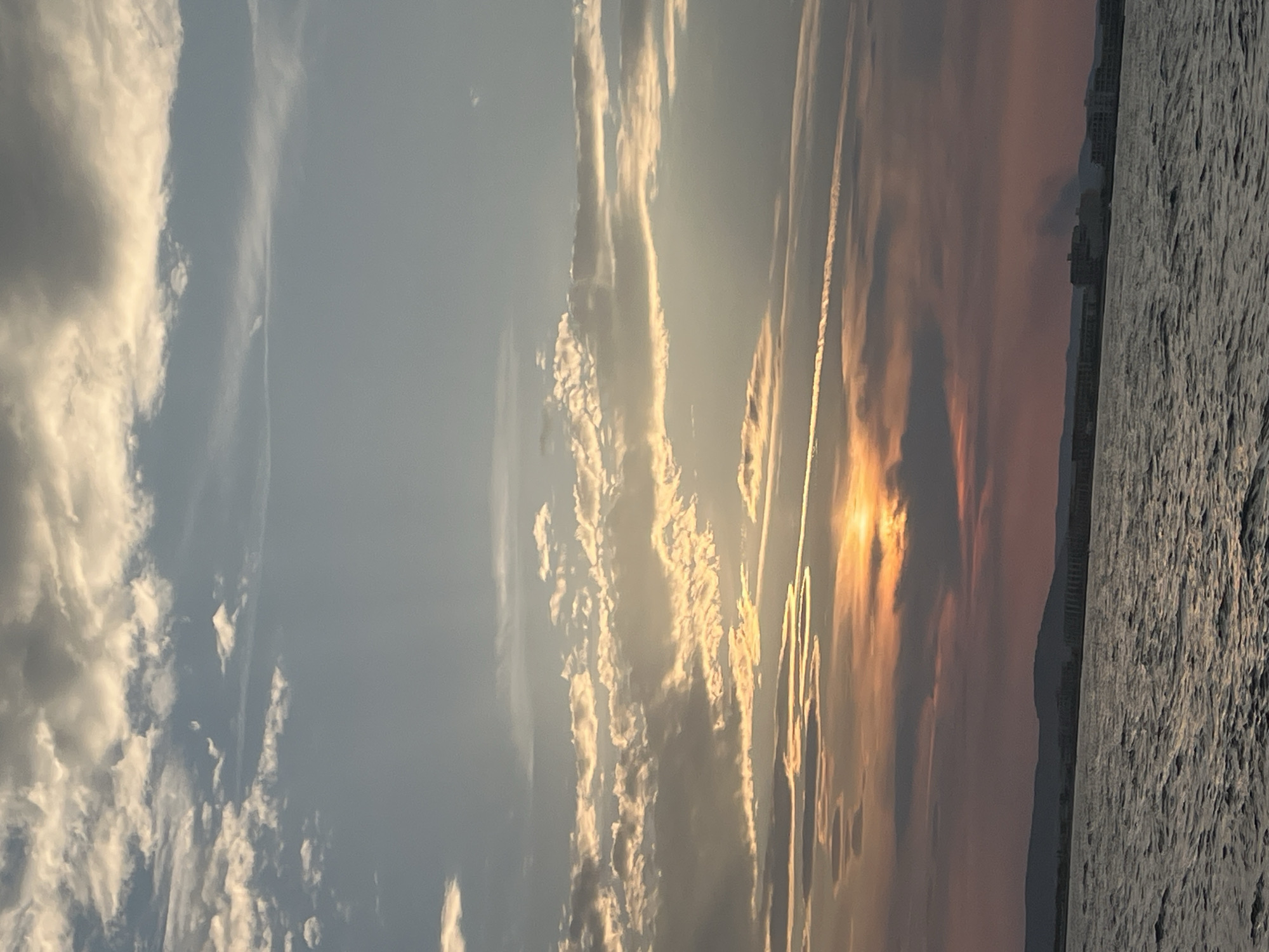 This image depicts a sunset over the sea, viewed from a body of water. The sky is filled with a variety of clouds, ranging from wispy cirrus to denser formations. The sun is partially obscured by clouds, casting warm hues of orange, pink, and yellow across the sky and illuminating the edges of some clouds. In the distance, a shoreline with a town or city is visible, characterized by a low-lying urban landscape backed by rolling hills. The water in the foreground is choppy, with small waves indicating a slight breeze. The overall scene is serene, capturing the atmospheric beauty of a sunset over a coastal environment.