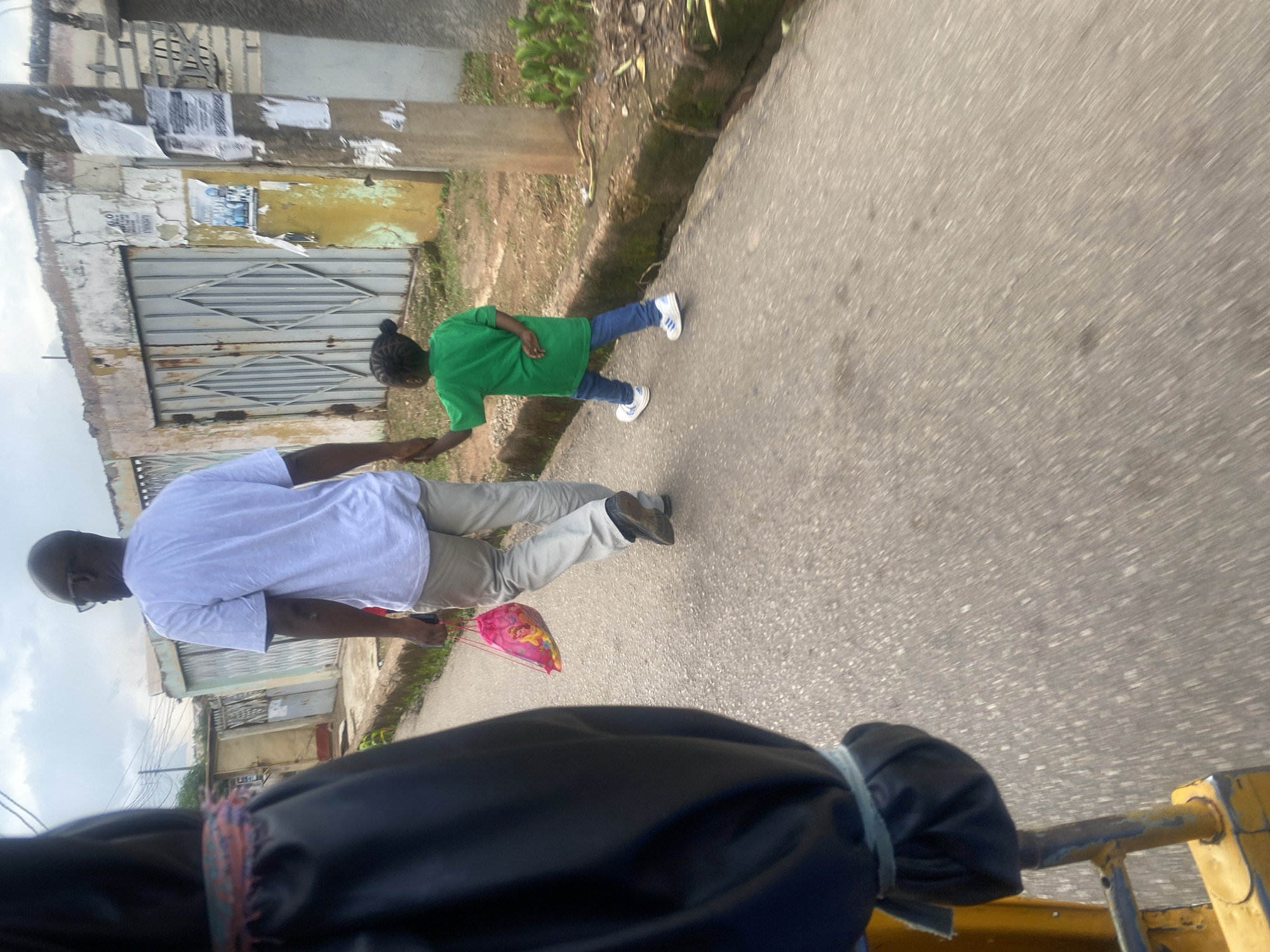 A man and a young child are walking hand-in-hand on a paved street in Jos, Nigeria. The man, wearing a white t-shirt and light-colored pants, carries a pink drawstring bag. The child, dressed in a green t-shirt and blue jeans, wears white athletic shoes. They are walking away from the camera, on the right side of the street. The background shows a weathered, pale yellow building with a large metal gate, and a utility pole covered in posters. The street is bordered by a raised concrete curb, with overgrown vegetation and dirt on the right side. The foreground is partially obscured by a black, tarp-like object with colorful trim, possibly part of a vehicle like a motorcycle taxi or rickshaw, suggesting the photo was taken from a moving vehicle. The sky is overcast, with visible clouds.