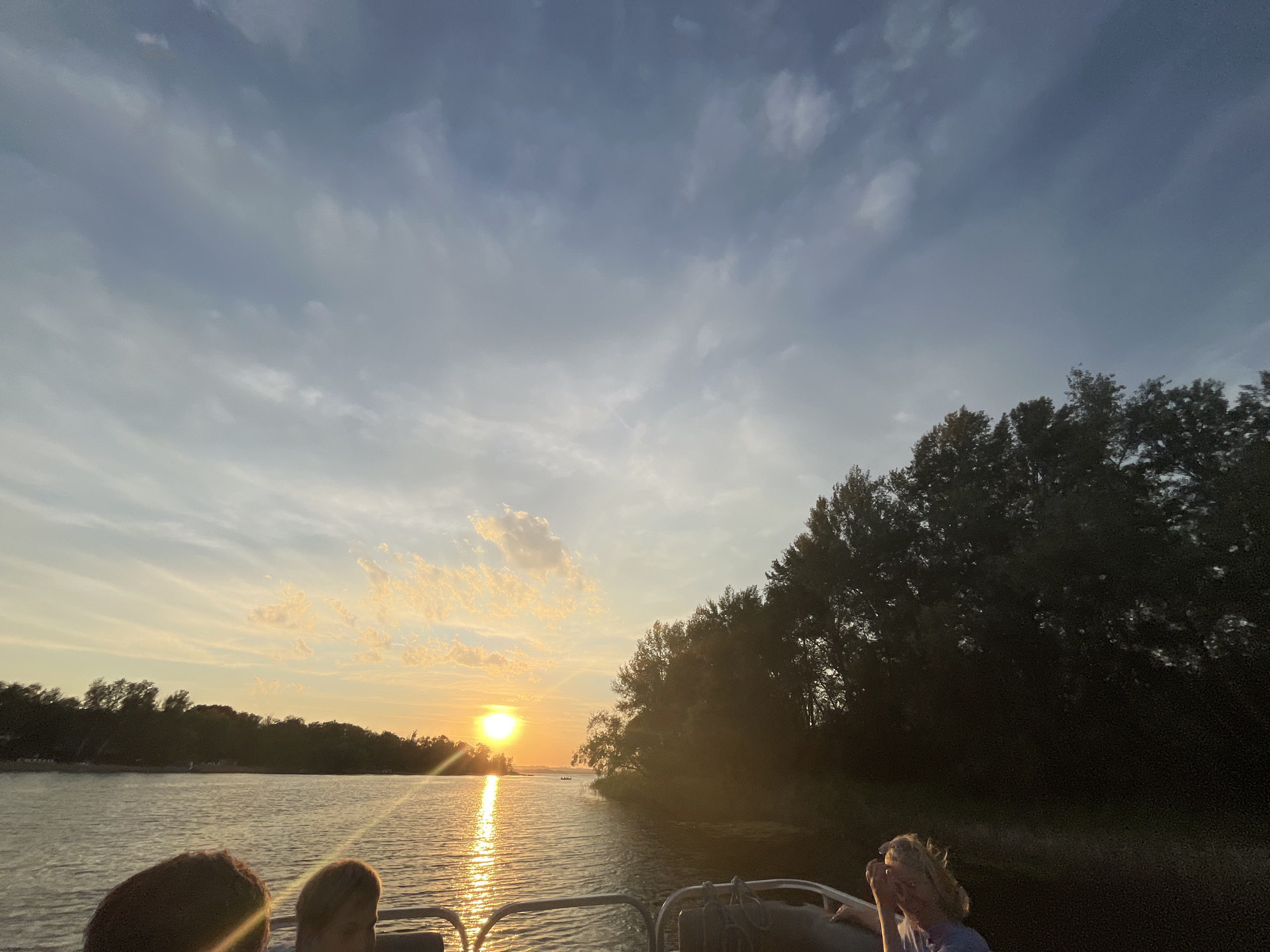 The image depicts a scenic sunset over a body of water, likely a river or lake, with a treeline on the right bank and a distant shoreline on the left. The sky is filled with a mix of clouds, with the setting sun casting a golden glow across the water. In the foreground, the edge of a boat is visible, with the heads of at least three people in profile. One person on the right, with blonde hair, appears to be looking down and touching their face. The overall atmosphere is serene, suggesting a leisurely boat ride during a summer evening. The location is specified as Shumeika, Russia.