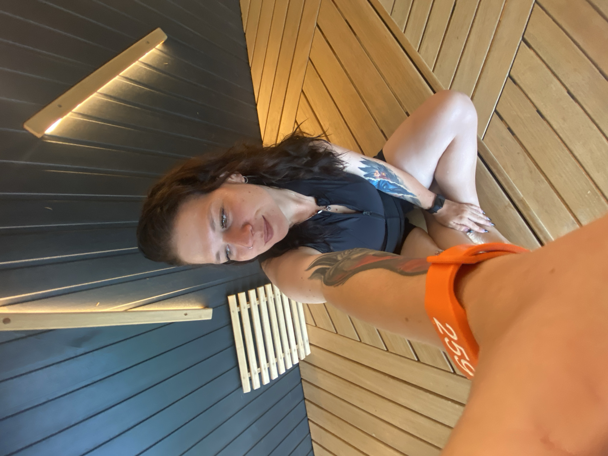 In a sauna, a woman with dark wavy hair and tattoos on her arms sits with her legs crossed. She wears a black zippered top. An orange wristband with the number "25g" is visible on her arm. The sauna features dark blue wooden walls and a slatted wooden ceiling with integrated lighting. The floor is made of light-colored wooden planks arranged in a chevron pattern. A small slatted wooden rack is attached to the dark blue wall. The lighting suggests it might be daytime, though the indoor setting makes it difficult to determine the exact time.