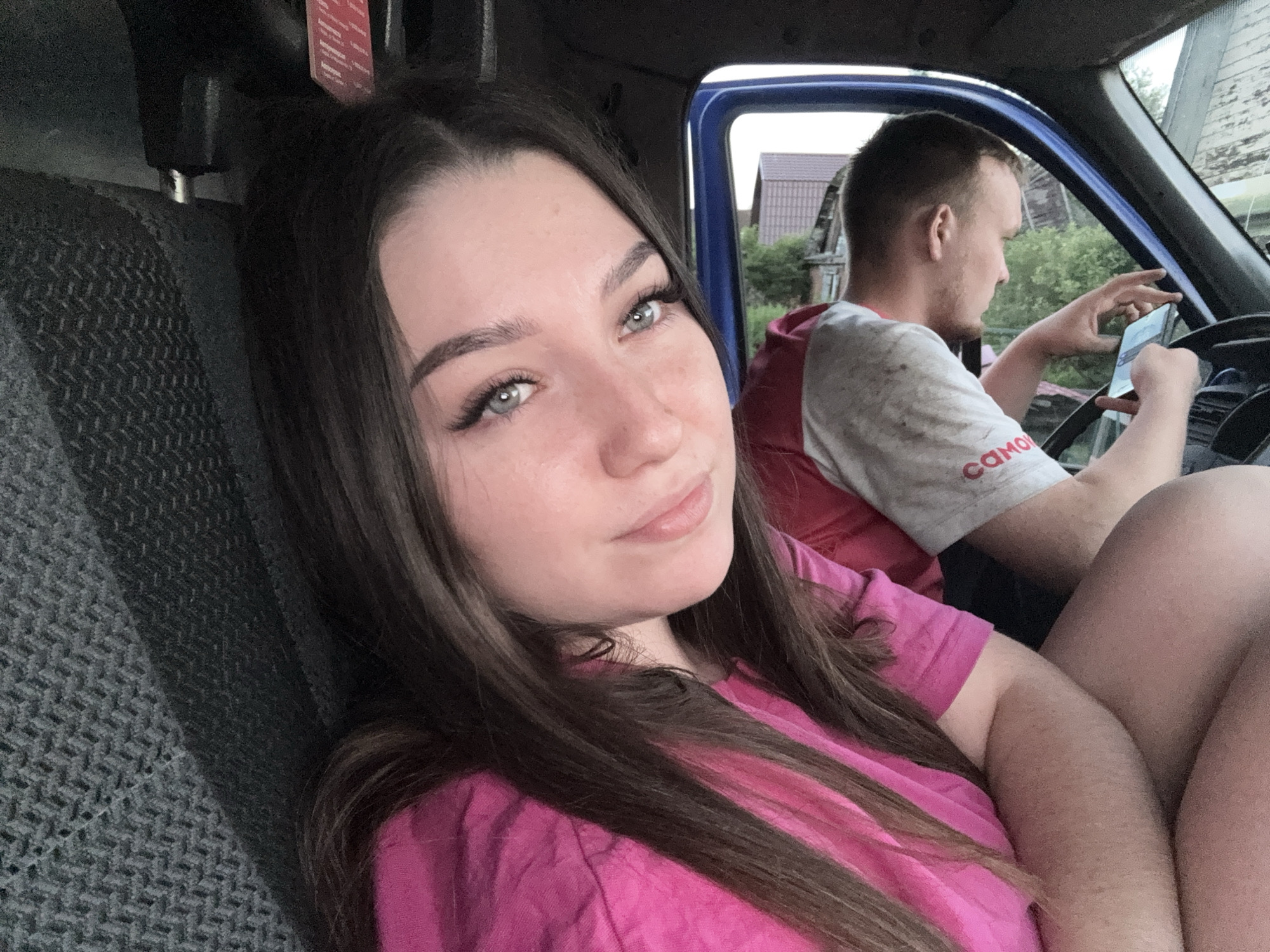 The image shows two people inside a vehicle, possibly a truck or van. In the foreground, a young woman with long brown hair and striking blue-green eyes is looking at the camera with a slight smile. She is wearing a bright pink t-shirt. To her right, in the background, a young man is visible, facing away from the camera and looking down at a smartphone he is holding. He is wearing a light-colored t-shirt with a red trim and some text on the sleeve. The interior of the vehicle appears somewhat utilitarian, with a textured dark gray seat visible on the left. Through the vehicle's blue door frame and window, a rural landscape with houses and greenery can be seen, suggesting an outdoor setting. The lighting indicates it is daytime, possibly late afternoon or early evening. The overall mood is casual and relaxed. There is text on the man's shirt sleeve that reads "САМО".