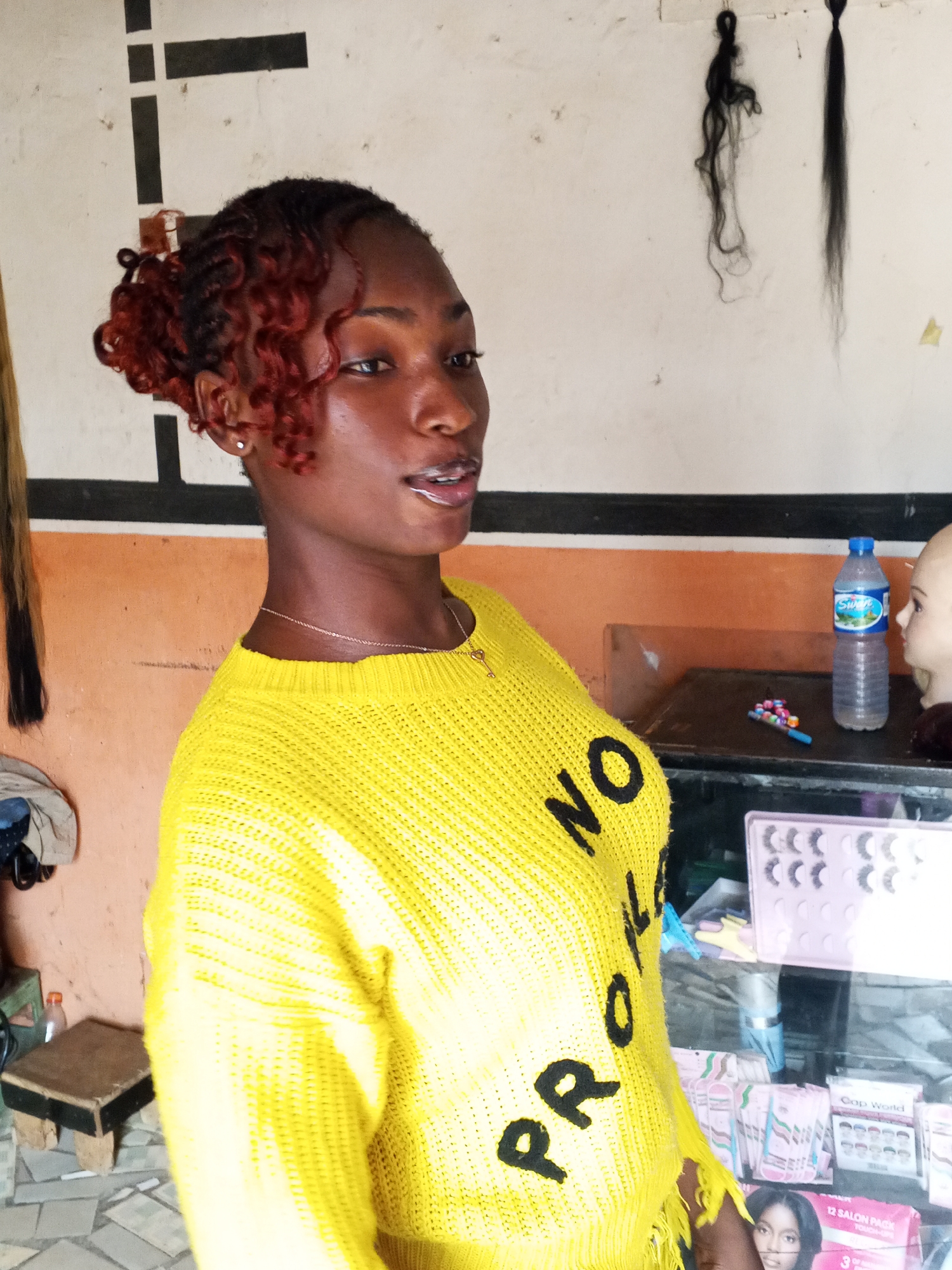 A young, dark-skinned woman stands in what appears to be a hair salon or beauty supply store. She is wearing a bright yellow sweater with black lettering that reads "NO PRO" and a thin gold chain. Her hair is styled in red-toned braids, with some styled to frame her face. She is facing slightly to the left of the frame. Behind her, the wall is divided horizontally: the upper portion is white with a series of black lines, while the lower portion is painted a reddish-orange color. Hanging from the ceiling are several bundles of artificial hair. To her right, a glass-fronted display case is filled with beauty products such as false eyelashes and hair products. A water bottle and mannequin head are visible on the countertop behind the display case. A small stool is present in the left foreground. The location is Gwong, Nigeria.