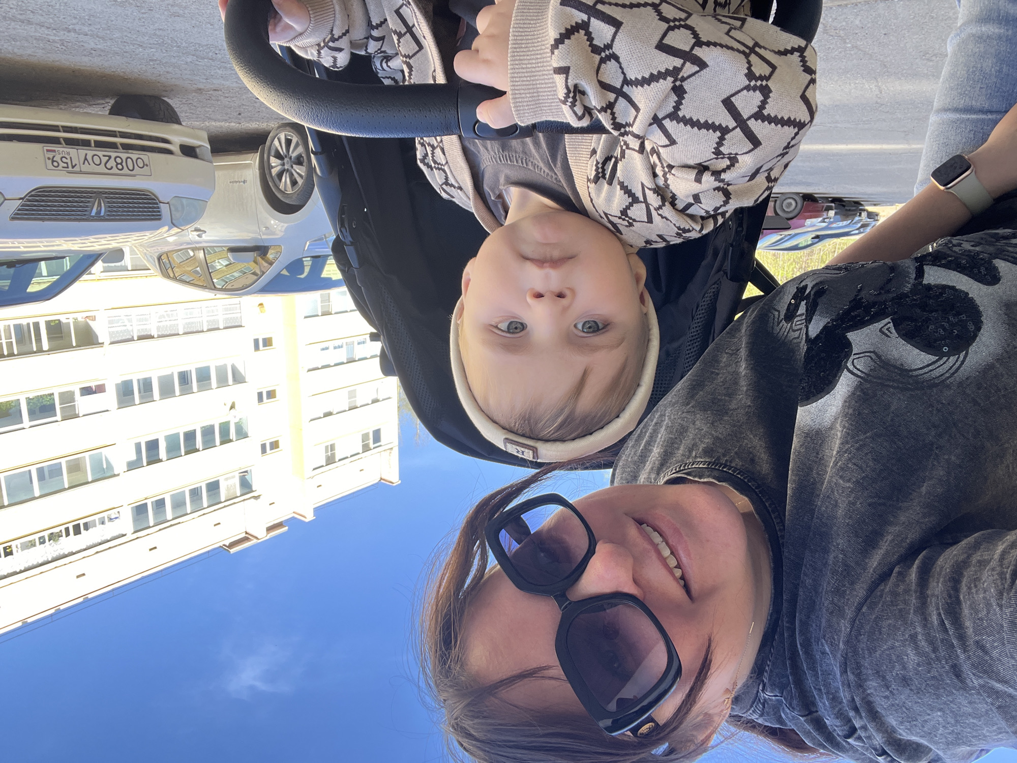 A woman and a baby are the main subjects of this outdoor shot. The woman is positioned closer to the camera and is wearing large, dark-rimmed sunglasses, a gray, textured jacket, and an Apple Watch on her wrist. She is smiling slightly. The baby is seated in a black stroller, wearing a light-colored patterned sweater and a matching headband. The baby has light skin and blue eyes and is looking directly at the camera.
The background features a multi-story beige building with numerous windows and a blue sky overhead. To the left, part of a white car is visible, with a license plate that reads "O082OY 159 RUS." Other cars can be seen parked further back. The photograph appears to be taken in Noviy, Russia, based on the car's license plate region code.