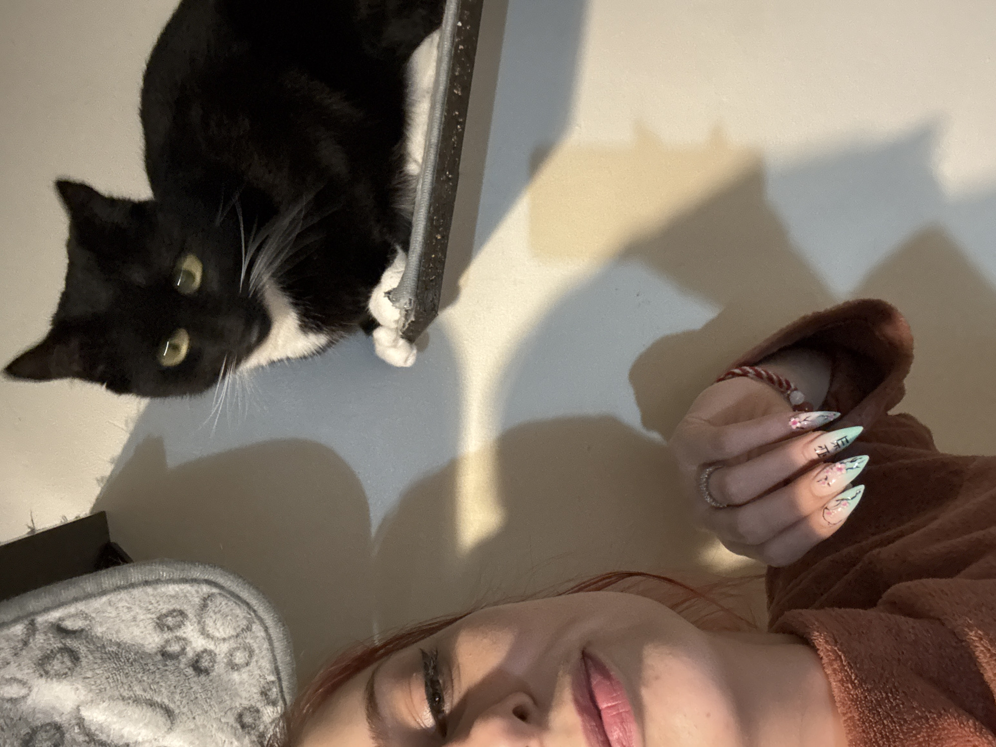 The image depicts a person lying down with a black and white cat positioned at the top of the frame. The cat has yellow eyes and white whiskers, and appears to be lying on a ledge or surface. The person's face is visible, and they have pink lipstick and reddish hair. Their hand is also visible, with elaborately decorated nails. There is a fuzzy grey object at the bottom left of the frame. The background is a plain wall. There is no clear evidence to ascertain the location as Desborough, United Kingdom.