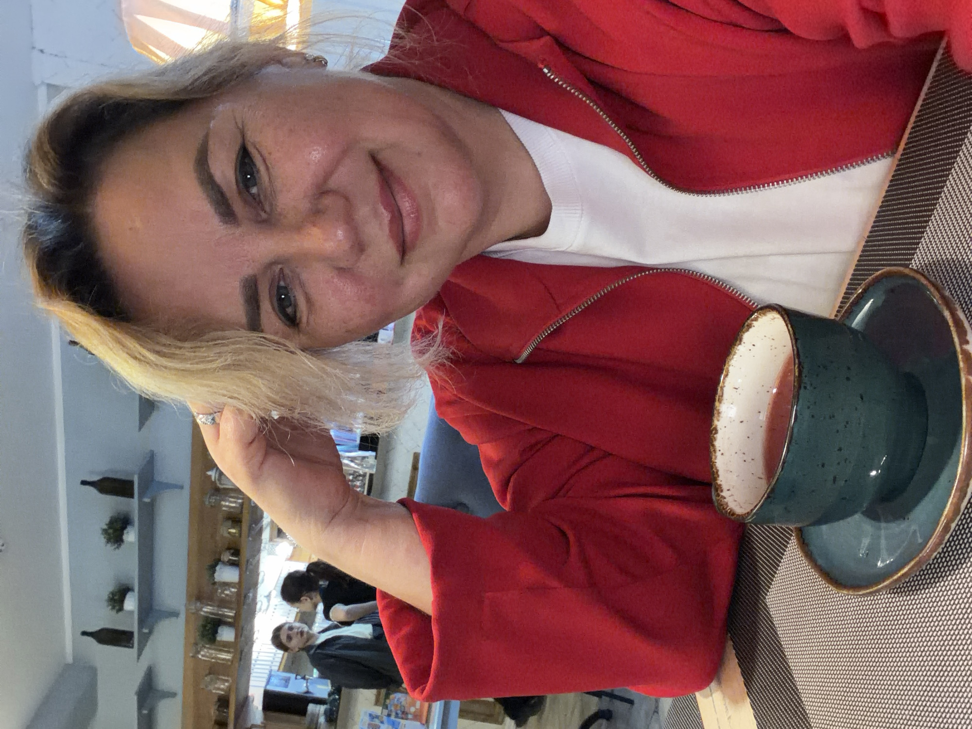 The image features a woman with light blonde hair and a red hooded jacket. She is smiling slightly and appears to be holding up her hand near her face. There is a cup and saucer on the table in front of her, suggesting she is having a drink. In the background, there is a blurred view of other people and shelves with decorative items.
The location appears to be a cafe or restaurant in Leningradskaya, Russia. The decor suggests an establishment in a modern style. The presence of other patrons indicates that the business is open and occupied.