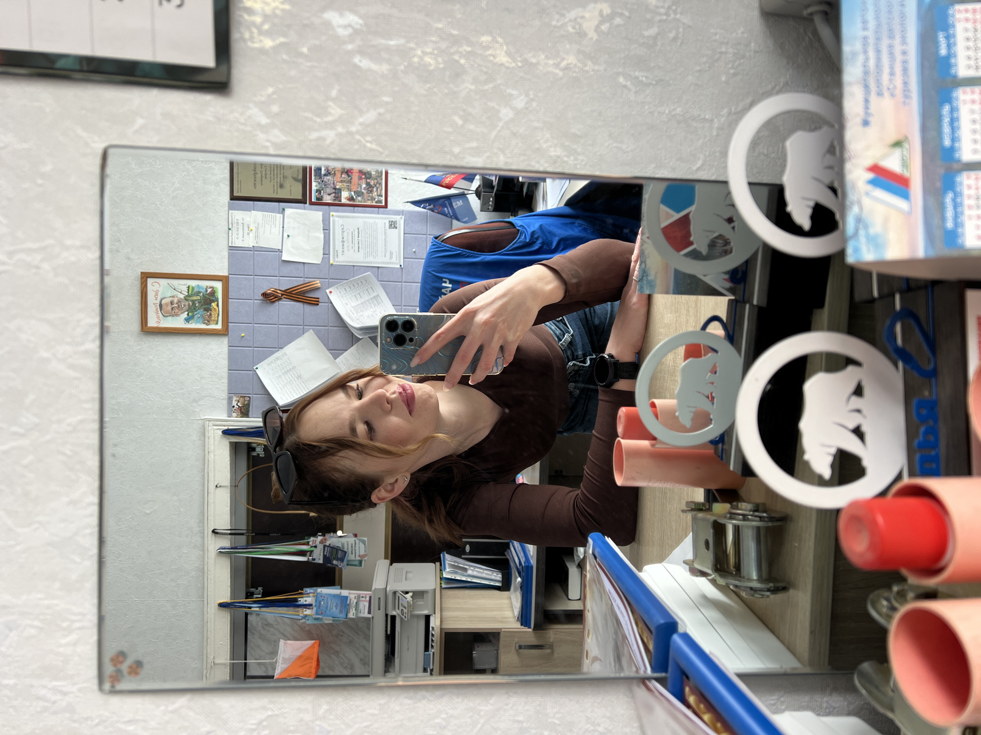 The image shows a reflection in a mirror. A woman is taking a selfie with a smartphone in the reflection. The mirror hangs in an office in Chaikovsky, Russia, as indicated by items on the wall and desk, including a Russian flag. The woman is wearing a brown long-sleeved shirt and jeans, and is wearing a watch on her left wrist. Sunglasses are perched on top of her head. In the background of the reflection are office items such as documents, calendars, and a blue vest. The room appears to be well-lit. The reflection also includes a bulletin board with photographs, certificates, and awards, and there is a picture of a man in military uniform on the wall. Objects on a desk near the mirror include paper rolls and pins with bear logos.