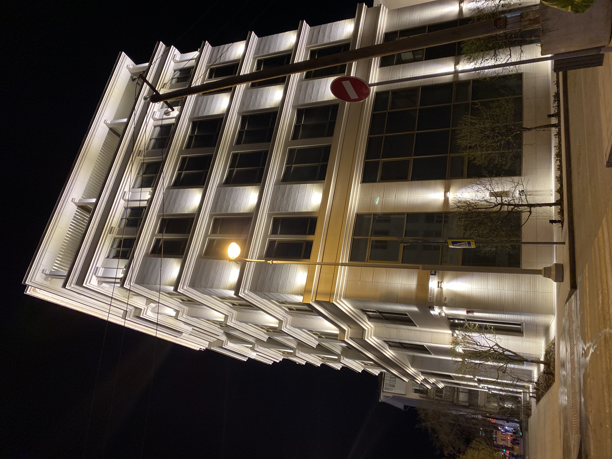 The image captures a nighttime view of a modern building in Yekaterinburg, Russia. The building, illuminated by external lights, features a multi-story facade with recessed windows and vertical architectural elements. A street sign indicates the address as Ulitsa Pushkina, 30. Overhead utility poles and streetlights are visible. A "no entry" traffic sign is mounted on one of the poles. Trees are planted along the building's facade. Sidewalks and possibly a road are visible on the right. The sky is dark and clear.