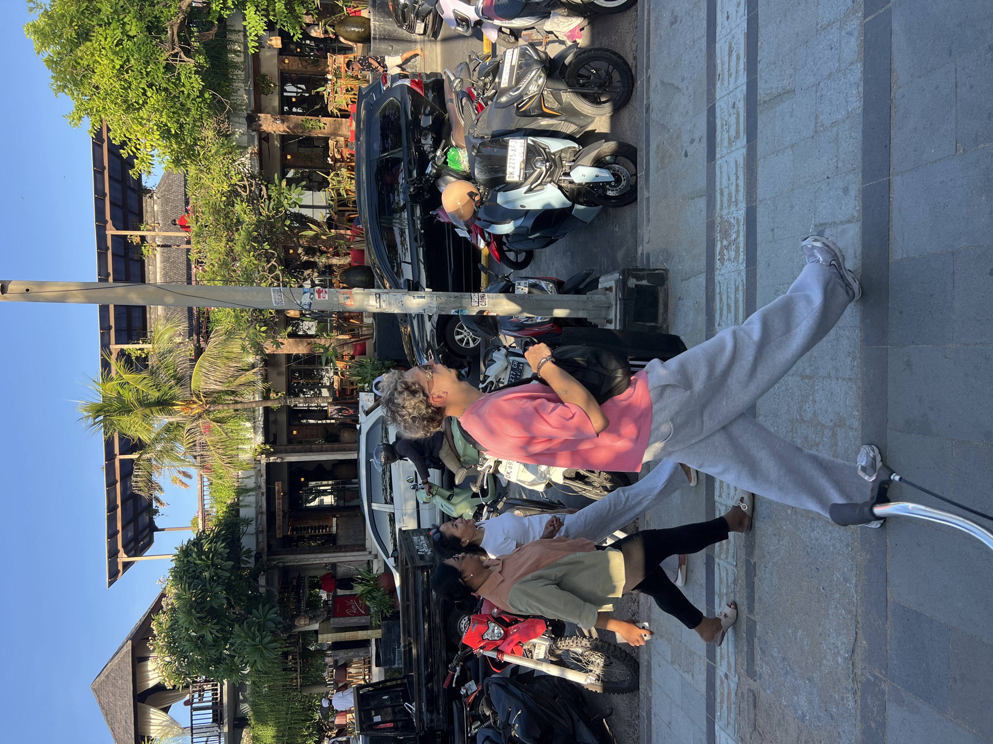 This is a wide shot of a street scene in Kuta, Indonesia, on a sunny day. The foreground shows the legs of a person walking, their bicycle handlebars visible. Three women walk together in the middle ground, two in darker clothing and one in a light brown and grey top. A man with curly blonde hair and a pink t-shirt walks in front of them. Behind them, a row of parked motorcycles is visible, with a black SUV and a white car further back. The background features a multi-story building with balconies and greenery, and trees. The building appears to be a restaurant or cafe with outdoor seating and thatched roofs on some levels. The overall atmosphere is that of a bustling commercial area.