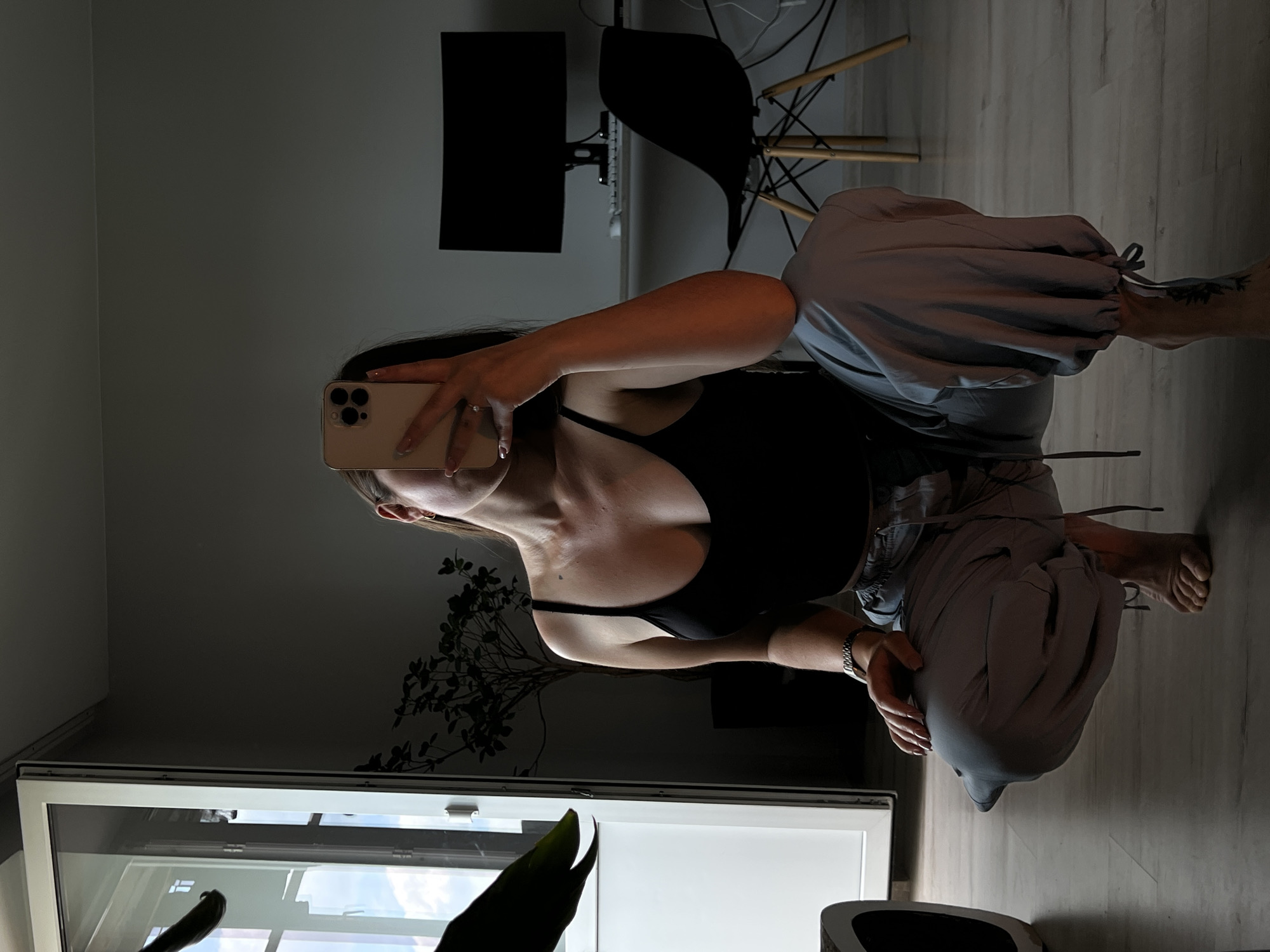 A woman is crouching and taking a selfie in a room. She is wearing a black tank top and light gray cargo pants. Her hair is dark brown and pulled back. She is holding a smartphone in her right hand, obscuring her face. In the background, there is a desk with a computer monitor and a chair. To the left, there is a large window with a white frame and a plant. The floor appears to be light-colored wood. The lighting is dim, with shadows cast across the room, suggesting it is either morning or evening. The image does not contain any specific information to place it in Ternopil, Ukraine.