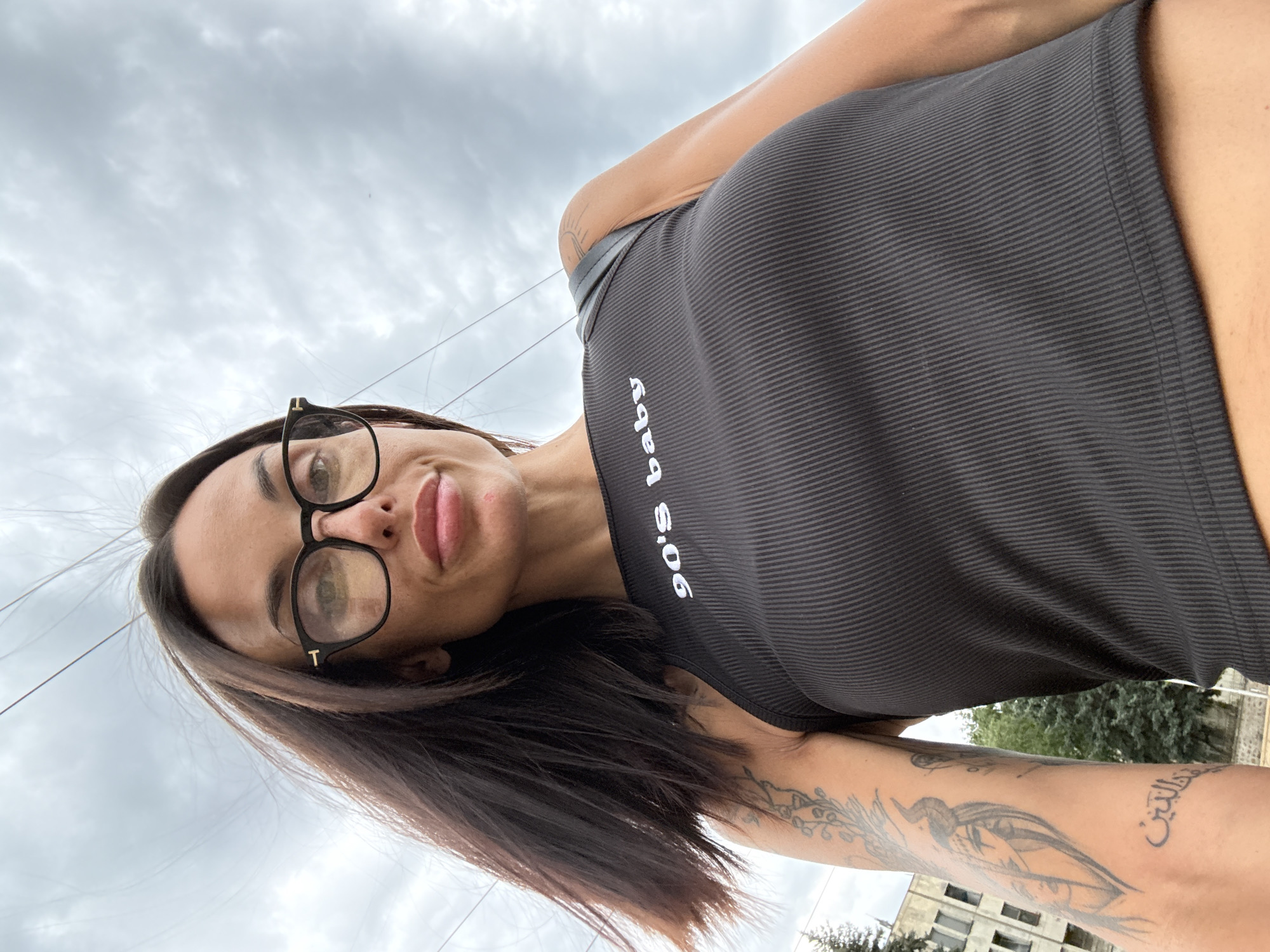 A woman with dark hair, wearing glasses and a black tank top with "90's baby" printed on it, is seen from a low angle against a cloudy sky. She has tattoos on her left arm. In the background, a building and trees are visible, suggesting an outdoor urban setting. The image appears to have been taken in the city of Pleven, Bulgaria.