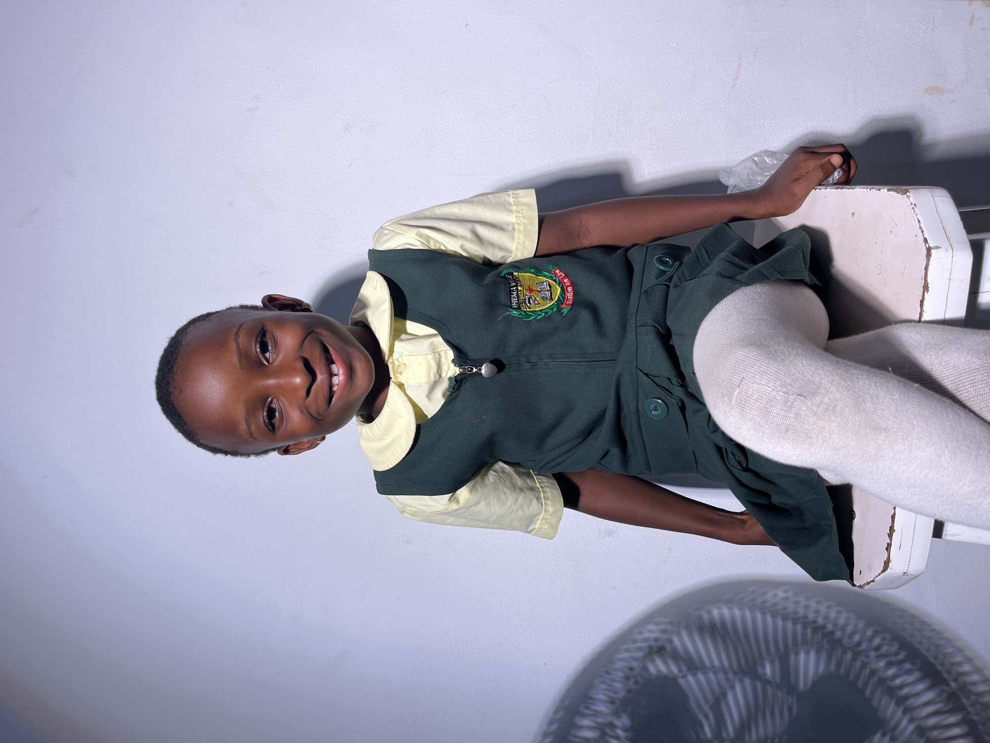 A young Black girl with a wide smile and dark, short hair sits facing the camera, her legs crossed at the knee. She is wearing a light yellow collared shirt under a dark green jumper dress. The dress has a school emblem embroidered on the left side of the chest. She is also wearing white tights. The girl is seated on a white, slightly distressed stool. To the left of the girl, a portion of a black fan with a white circular pattern is visible, casting a striped shadow on the wall behind her. The background is a plain, light-colored wall. The lighting is direct, creating some shadows. The image appears to be taken indoors, in an area that could be a school or home. The specific city is unknown.