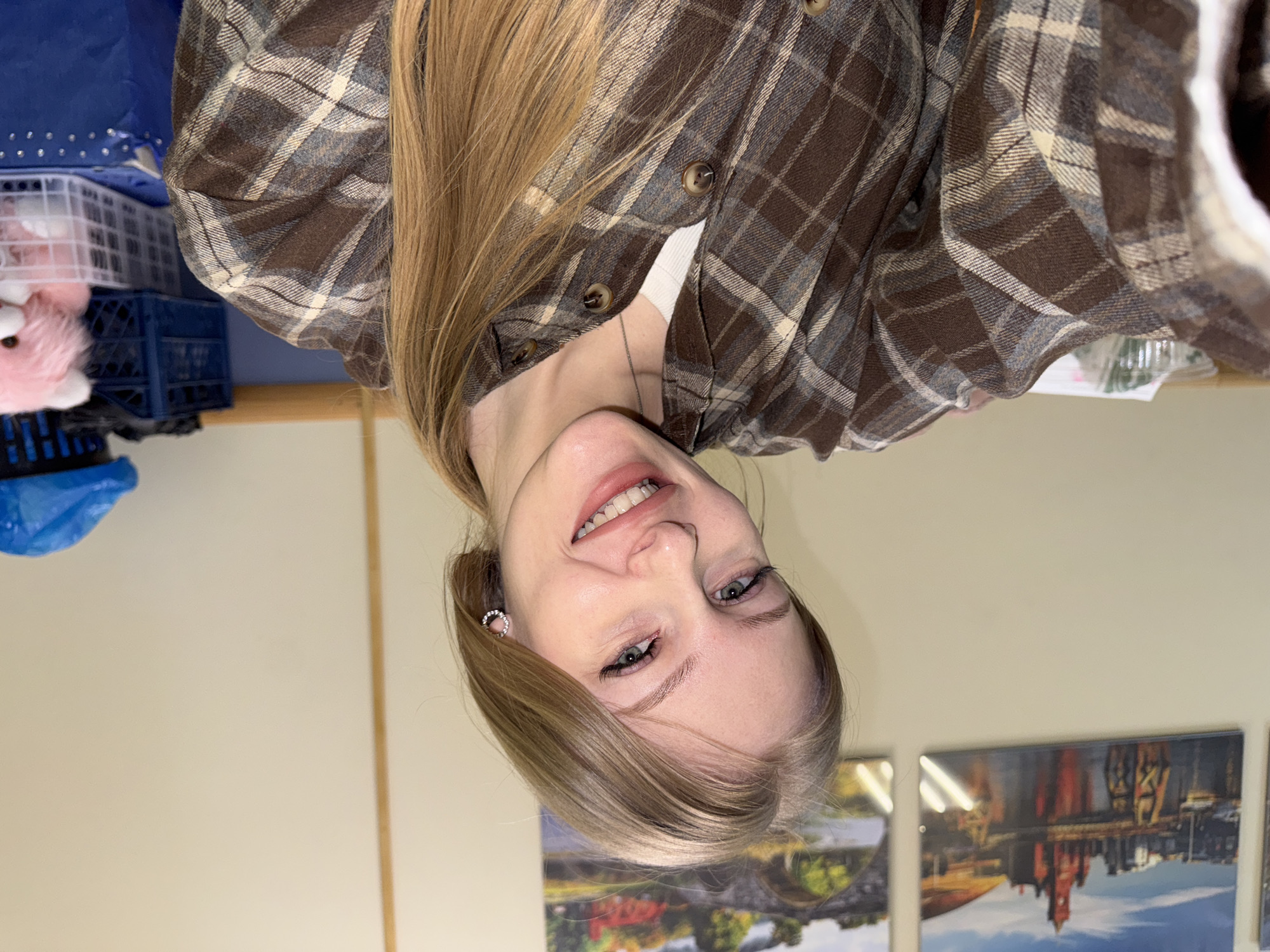 The image shows a blonde woman wearing a brown plaid shirt. The background is a light beige wall with three framed pictures. A blue storage container and a white basket are visible in the upper left corner. Without additional information or clues within the image, determining the exact location in Perm, Russia, is impossible.