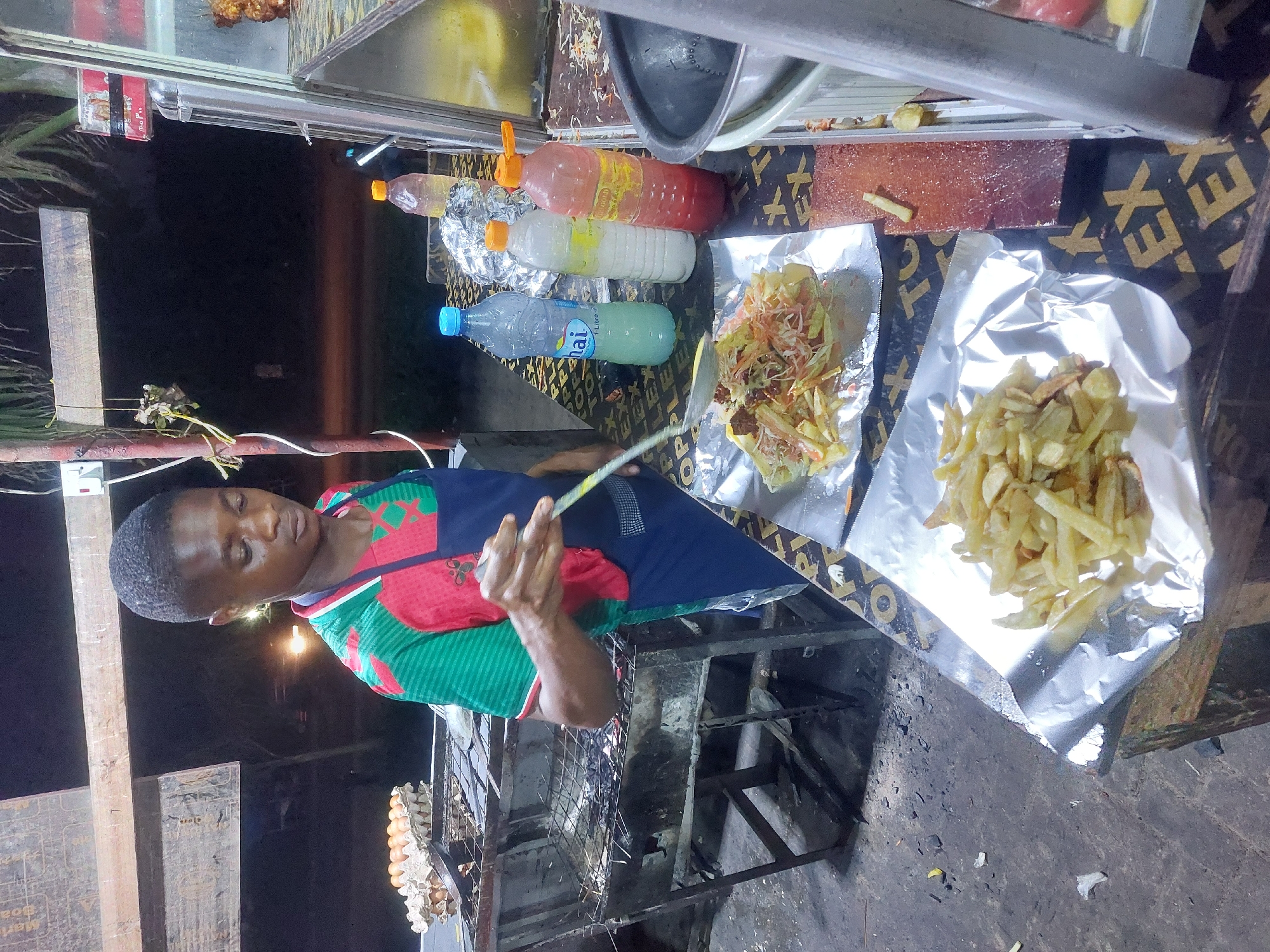 The image shows a street food vendor in Dar es-Salaam, Tanzania, preparing food. The vendor, a young man with short, dark hair, is wearing a red and green patterned shirt and a dark blue apron. He is holding a utensil in his right hand, presumably for cooking or preparing food.
In front of him is a makeshift food stall with several items. There are bottles of different sauces and drinks, including one labeled "Nai," a brand of bottled water, and another with a red sauce. Two foil-lined trays hold cooked food: one contains French fries, and the other appears to be a mix of vegetables and other ingredients, possibly a salad or a prepared dish of some kind.
Below the countertop, a grill sits over what appears to be a charcoal or wood fire, judging by the darkened metal. To the left of the grill, a carton of eggs is visible. The stall is placed on a surface with patterned lettering. The ground is visibly dirty. The environment suggests a typical bustling street-food scene in the city.