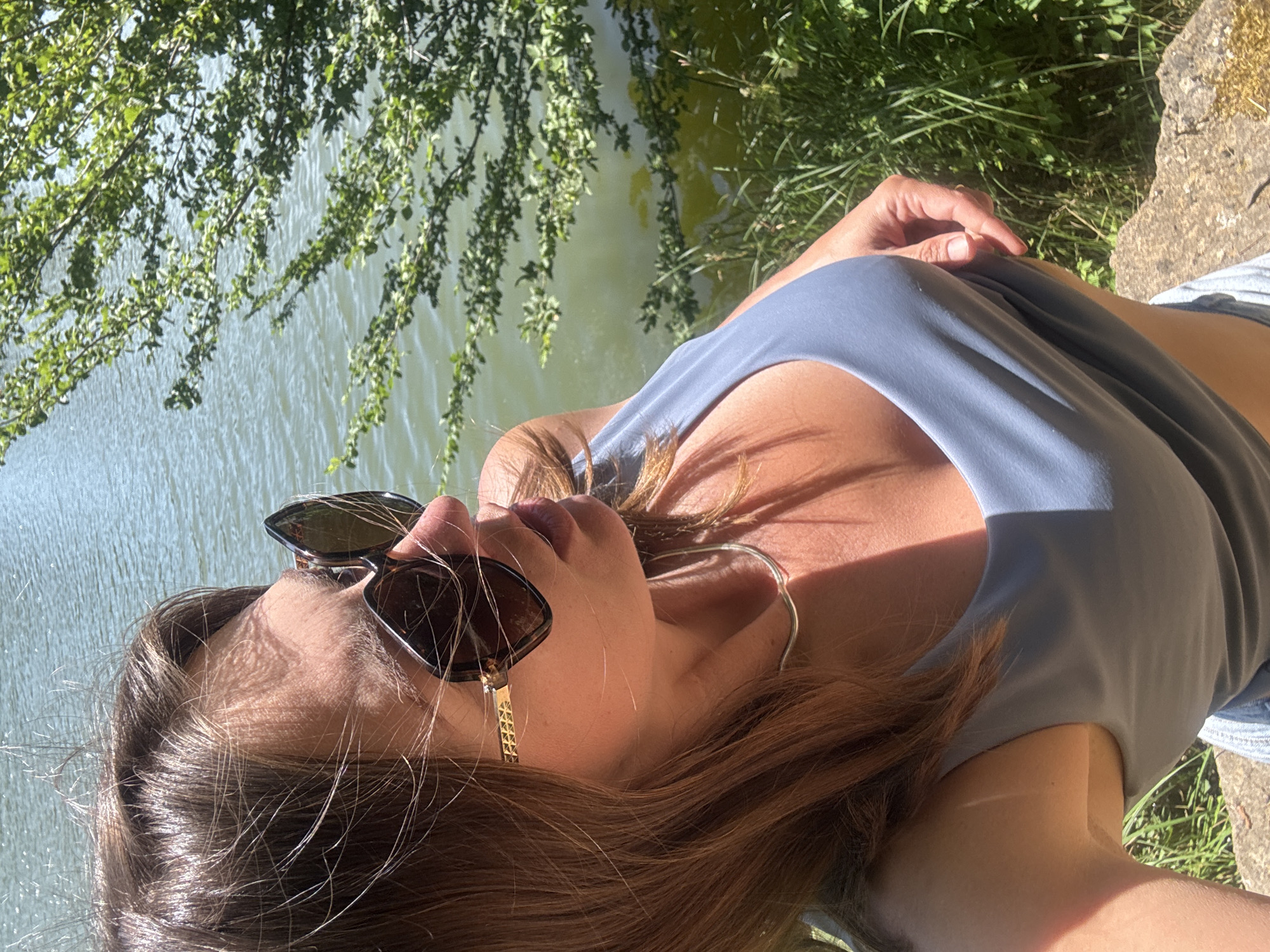 The image shows a woman with shoulder-length brown hair and sunglasses, wearing a blue tank top and denim shorts. She is standing outdoors, with a body of water behind her and green foliage to the right. She is holding up the bottom of her tank top with her right hand, exposing her midriff. The setting appears to be a park or natural area, possibly near a lake or river. The image does not contain information that allows for identification of the specific location within Ravilloles, France.