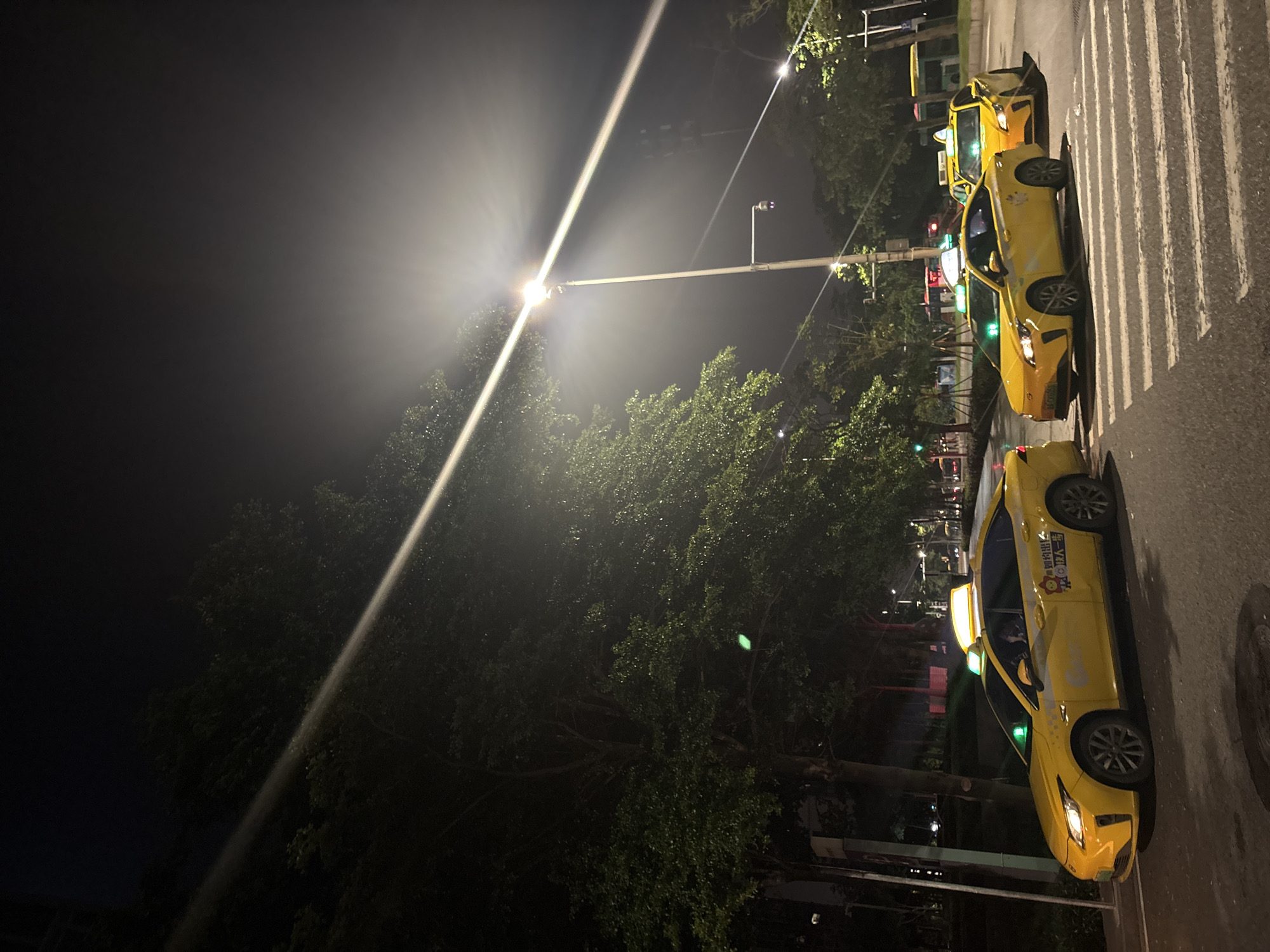 The image shows a nighttime scene in Haizhu District, China, featuring several yellow taxis lined up on a street with a pedestrian crossing. The sky is dark, and a bright light source, possibly a street lamp or a spotlight, casts a strong beam across the scene, creating a hazy effect and illuminating the trees and taxis. The weather appears to be clear, with no visible rain or fog.
The primary subjects are the yellow taxis, with at least three clearly visible, parked on the road. The closest taxi is a bright yellow sedan with a light on its roof and markings on its doors, including some text in what appears to be Chinese characters and a cartoonish graphic. The other two taxis are further down the street, also bright yellow. The scene includes a pedestrian crossing marked with white stripes on the asphalt road. Trees with green foliage line the side of the road, and streetlights and traffic signals are visible in the background.
The time of day is clearly night, indicated by the dark sky and artificial lighting. The overall mood is calm and quiet, typical of a nighttime street scene. There are no people visibly interacting with the taxis or the environment, suggesting they are parked or waiting. The presence of multiple taxis and a pedestrian crossing hints at a public area, possibly near a transportation hub or a busy street. Some visible text on the taxi includes Chinese characters and what appears to be a logo or advertisement.