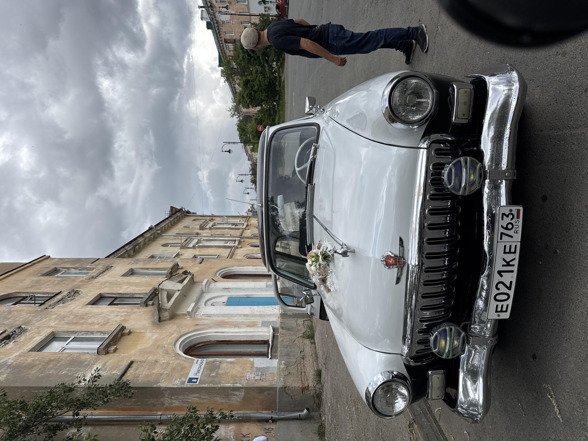 A white and black vintage GAZ-21 "Volga" car, adorned with a small floral bouquet and lace on its hood, is parked on a paved street. The car's license plate, E021KE 763 RUS, is visible on the front bumper. The front passenger door is ajar, and a white steering wheel is discernible through the windshield. Two auxiliary circular lights are mounted on the front bumper.
To the left of the car, an aged, light brown multi-story building with peeling paint and architectural details, including arched and rectangular windows, lines the street. A blue door is visible within the building's facade further down. A blue street sign, displaying "8 Никонова" (8 Nikonova), is affixed to this building. Green foliage partially obscures the building's far left side.
A man, wearing a black t-shirt, blue jeans, and a light-colored cap, stands to the right of the car, facing away from the camera and looking towards the distant background. The sky is overcast with heavy grey clouds. In the background, an urban streetscape includes additional buildings, trees, streetlights, and a red car parked further down the road. A "No Entry" sign is also visible in the distance. The scene is located on Nikonova Street in the city of Yaroslavl, Russia.