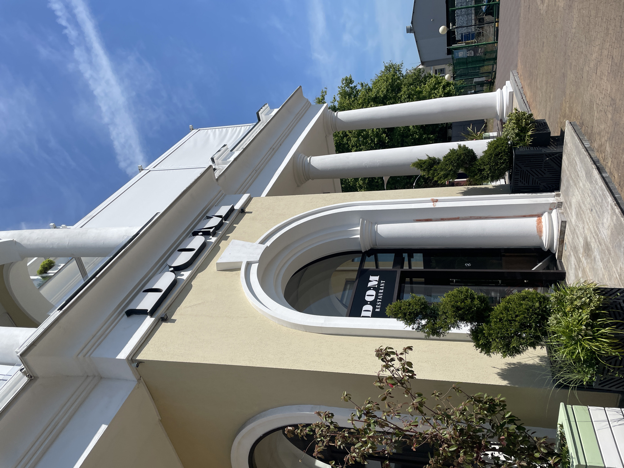 This full-frame, eye-level outdoor shot depicts the facade of a light yellow stucco building with white classical architectural elements on a sunny day in Sochi, Russia. The building features multiple white columns with Doric or Tuscan capitals supporting an extensive white cornice, and large arched window/doorways. The main entrance, centrally located, is an arched glass door displaying "DOM RESTAURANT" in white lettering on a dark background. Above the entrance, large, dark, raised letters spell "DOM" on the building's facade.
In the foreground, various potted plants flank the entrance, including a small tree with reddish-green foliage in a light green and white striped planter to the left, and a spiral-shaped evergreen topiary along with other leafy green plants in black geometrically patterned planters closer to the door. The ground in front of the building is paved with reddish-brown bricks.
Beyond the columns to the right, lush green trees are visible. Further in the background, a light blue sky with a distinct, wispy white contrail spans the upper right corner. A glimpse of a gray building with a fence and what appears to be an industrial or construction area is visible in the far right background. No people are present in the frame. Shadows are prominent, indicating bright daylight, likely midday or early afternoon.