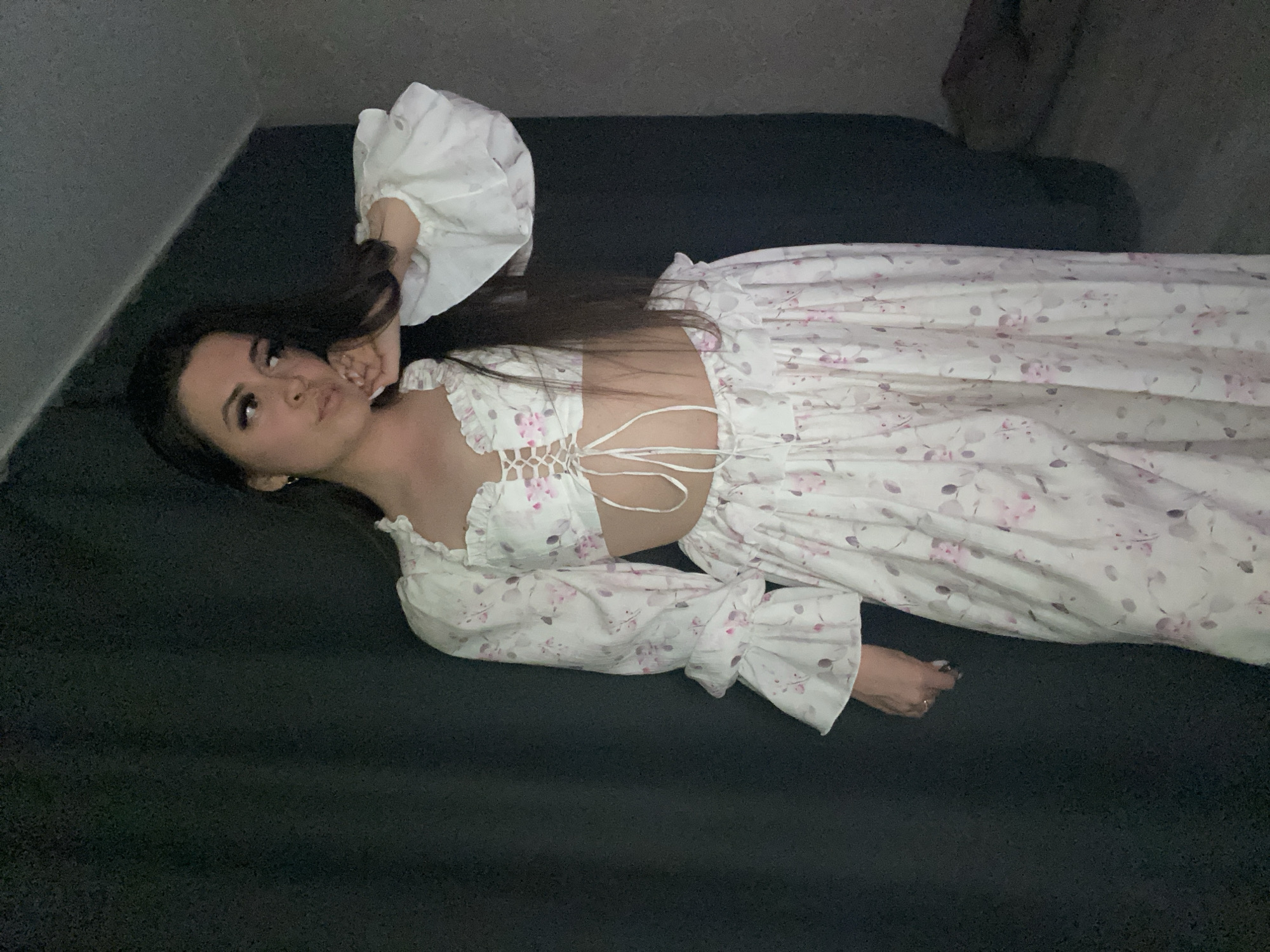 A woman lies on a dark-colored surface, possibly a bed or sofa, with her head near the upper left corner and her body extending diagonally toward the lower right. She is wearing a light-colored outfit with a floral pattern, consisting of a top that laces up the front and a long skirt or pants. The top has ruffled details around the edges and large, puffed sleeves. Her long, dark hair is spread out around her head. Her left hand is near her face, and her right hand is near the bottom edge of the frame. The background is a dimly lit wall. The location is Chaikovsky, Russia.