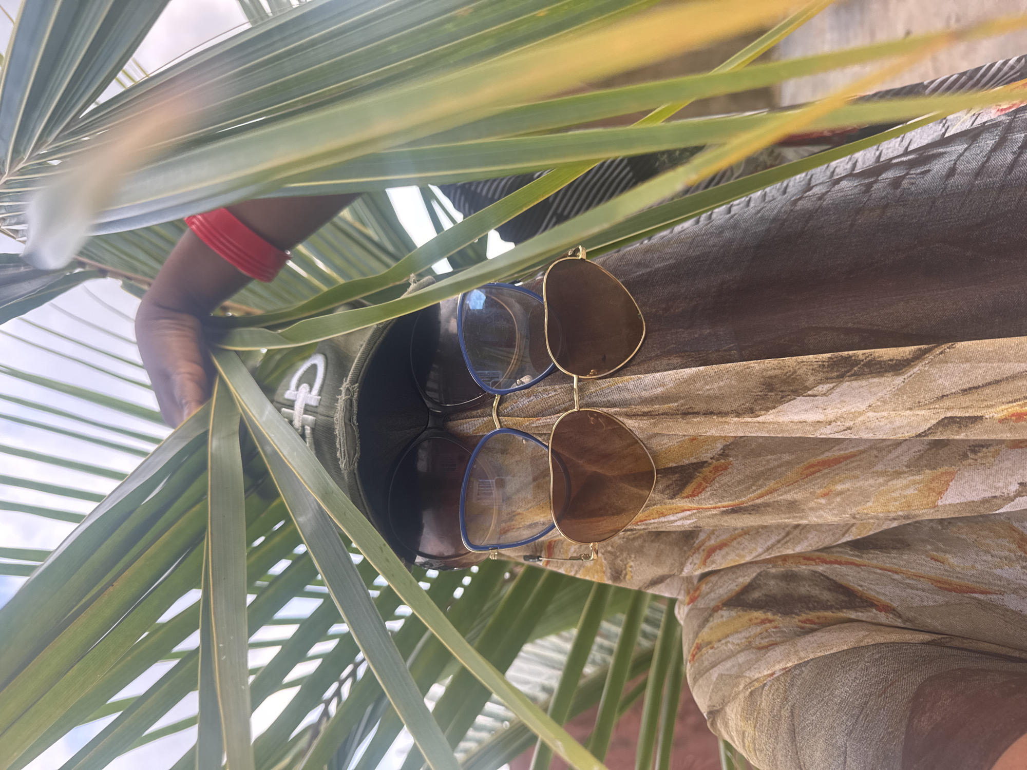 A person is partially obscured by palm fronds and is wearing a patterned fabric over their head and shoulders, a green cap, and two pairs of sunglasses, one over the other. The person has a red bracelet on their left wrist. The background appears to be a slightly out-of-focus outdoor environment with a hint of a building. The lighting suggests it is daytime. There is no specific indication of the location being in Jawu, Nigeria, or any other city.