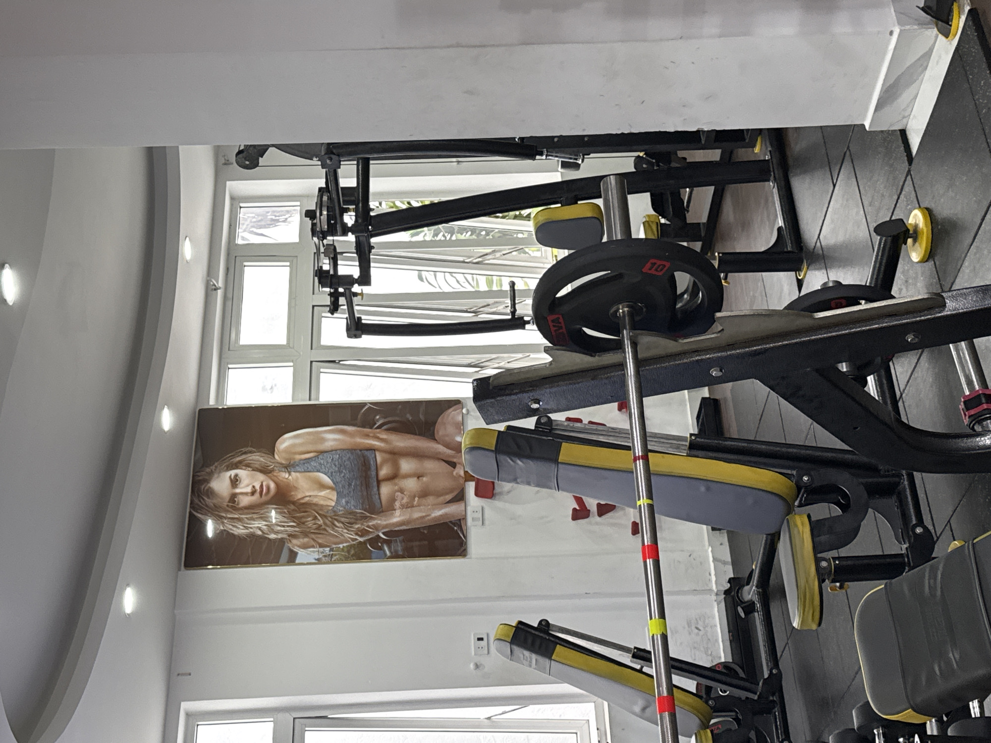 This image captures the interior of a gym in Da Lat, Vietnam, set up for weight training, with no people present. The scene is dominated by various exercise equipment, suggesting it is ready for use.
In the foreground, a prominent weight bench, likely part of a multi-gym machine, is visible with grey and yellow padded sections. A barbell, featuring a black 10kg weight plate with red numbering and a partially visible "NA" or similar text, rests on supports. Other black and yellow gym equipment, including additional benches or machine parts and weight plate storage, can be seen throughout the room. The floor is dark and tiled, likely rubber gym flooring.
On the left wall, a large framed poster depicts a muscular woman with long, wavy hair, wearing a grey sports bra, showcasing a strong physique with visible abdominal muscles. Her expression appears determined. Large windows are present in the background and near the poster, letting in natural light, suggesting it is daytime. Overhead, modern recessed ceiling lights illuminate the space. The walls and ceiling are white, with some architectural curves on the ceiling.