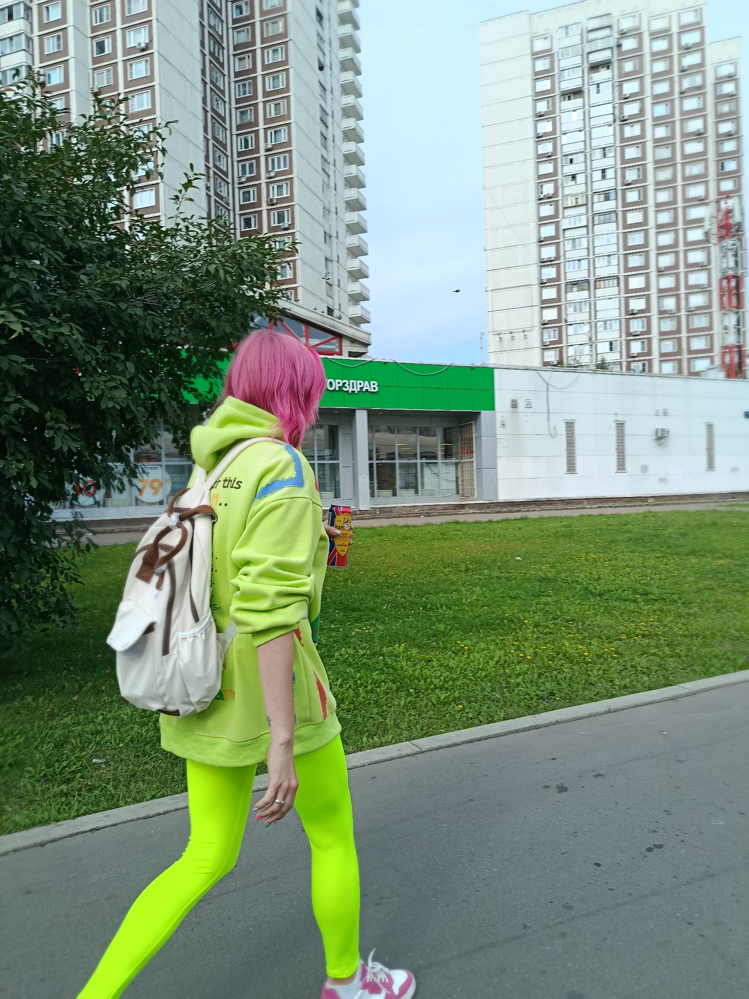 A person with bright pink hair walks on a sidewalk in Moscow, Russia, wearing a bright green hooded sweatshirt and bright green leggings. The person carries a white backpack and a red and white can in their right hand. In the background are tall apartment buildings and a green building with the Cyrillic word "ЗДОРОВЬЕ" (zdorovye) which translates to "Health". The area is covered in grass and trees.