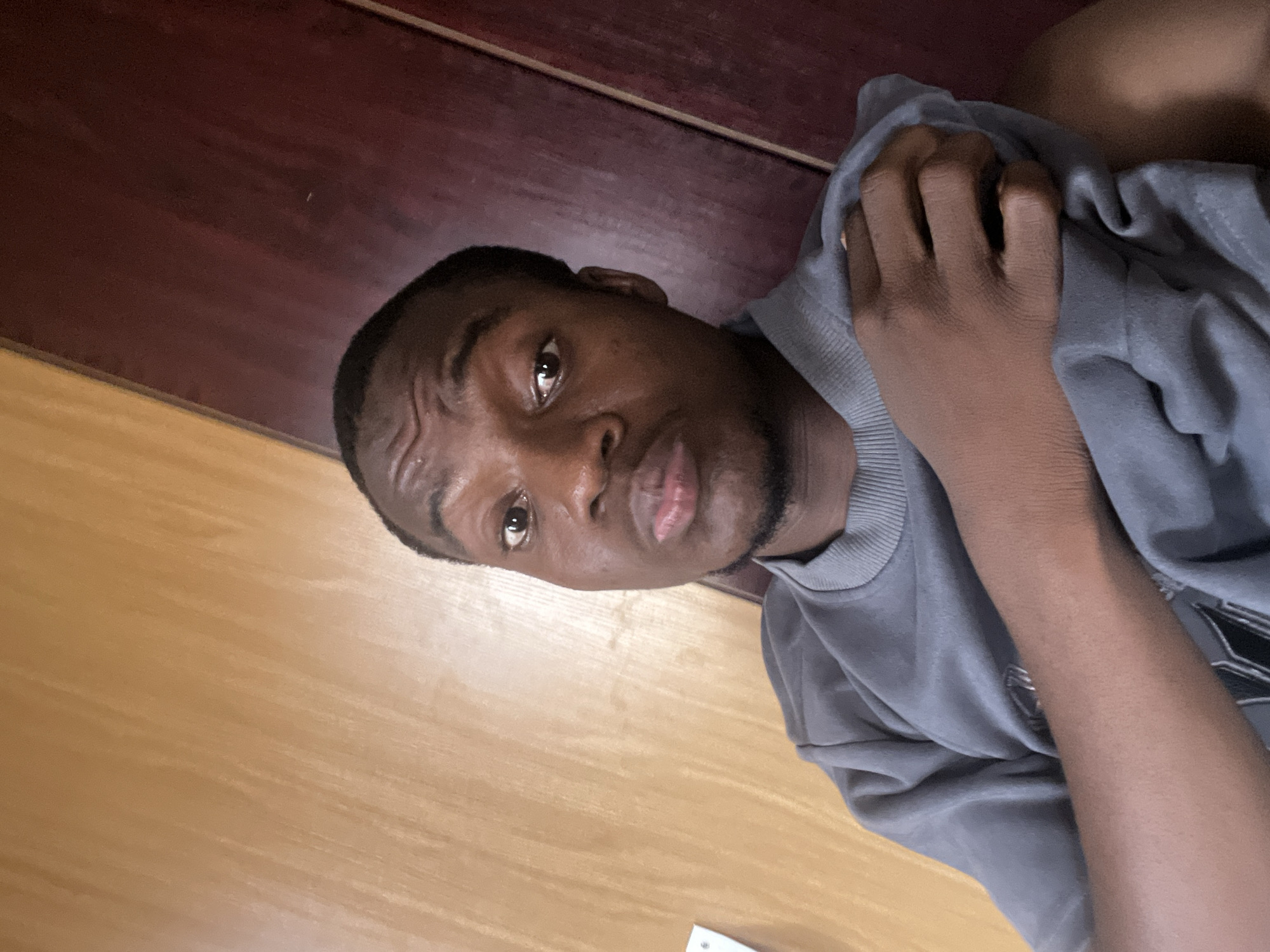 A close-up, slightly angled shot features a young man with dark skin and short hair. He is wearing a grey sweatshirt and is holding the sweatshirt with his left hand, positioned close to his face. His expression appears serious. The background reveals a wooden surface, possibly a floor or table, with a darker wooden surface partially visible. The location is presumed to be in Minna, Nigeria, but there are no visual cues to confirm this. No specific actions, interactions, or objects of forensic interest are readily apparent.