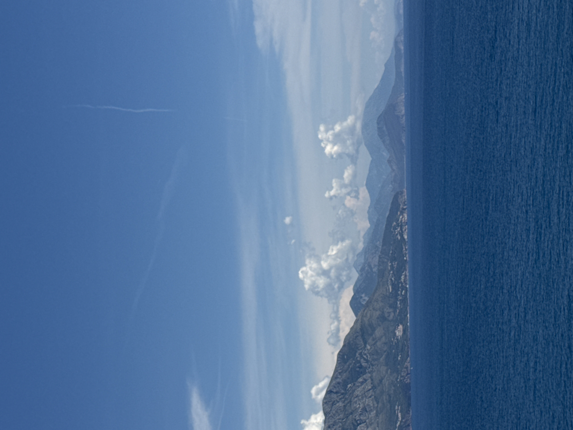 The image depicts a scenic coastal landscape under a bright blue sky with scattered clouds. In the foreground, a vast expanse of deep blue sea occupies the lower half of the frame. The sea surface exhibits a gentle ripple, indicating calm conditions.
In the background, a rugged mountain range stretches horizontally across the image, forming the coastline. The mountains are covered in vegetation, with some areas appearing rocky and bare. Along the lower slopes of the mountains, hints of human settlement are visible, suggesting a town or village nestled against the mountainside. The architectural style appears to be traditional, with buildings clustered together.
The sky above is a vibrant blue, characteristic of a sunny day. wispy white clouds are dispersed across the sky, with a notable condensation trail from an aircraft visible as a thin, vertical white line in the upper center of the image. The overall impression is one of natural beauty and a tranquil coastal environment. The location is identified as Dubraljevina, Montenegro.