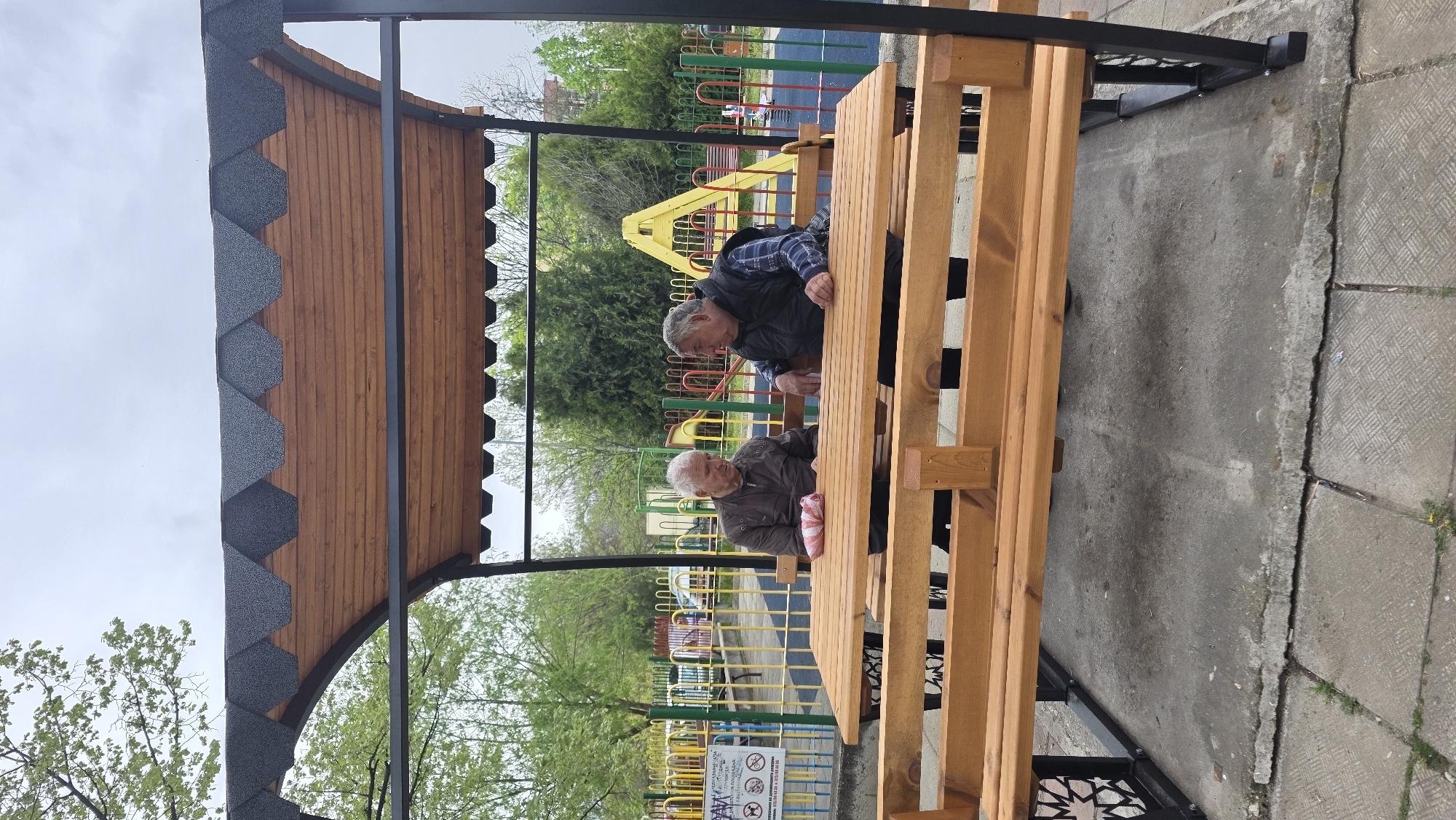 Two elderly men sit at a wooden picnic table under a shelter with a shingled roof in a park setting. One man wears a dark jacket and plaid shirt, while the other wears a brown jacket. A playground with colorful equipment is visible in the background, beyond a fence. The shelter has black metal supports and decorative cutouts. The ground is paved with concrete slabs. The scene appears to be in Blagoevgrad, Bulgaria.