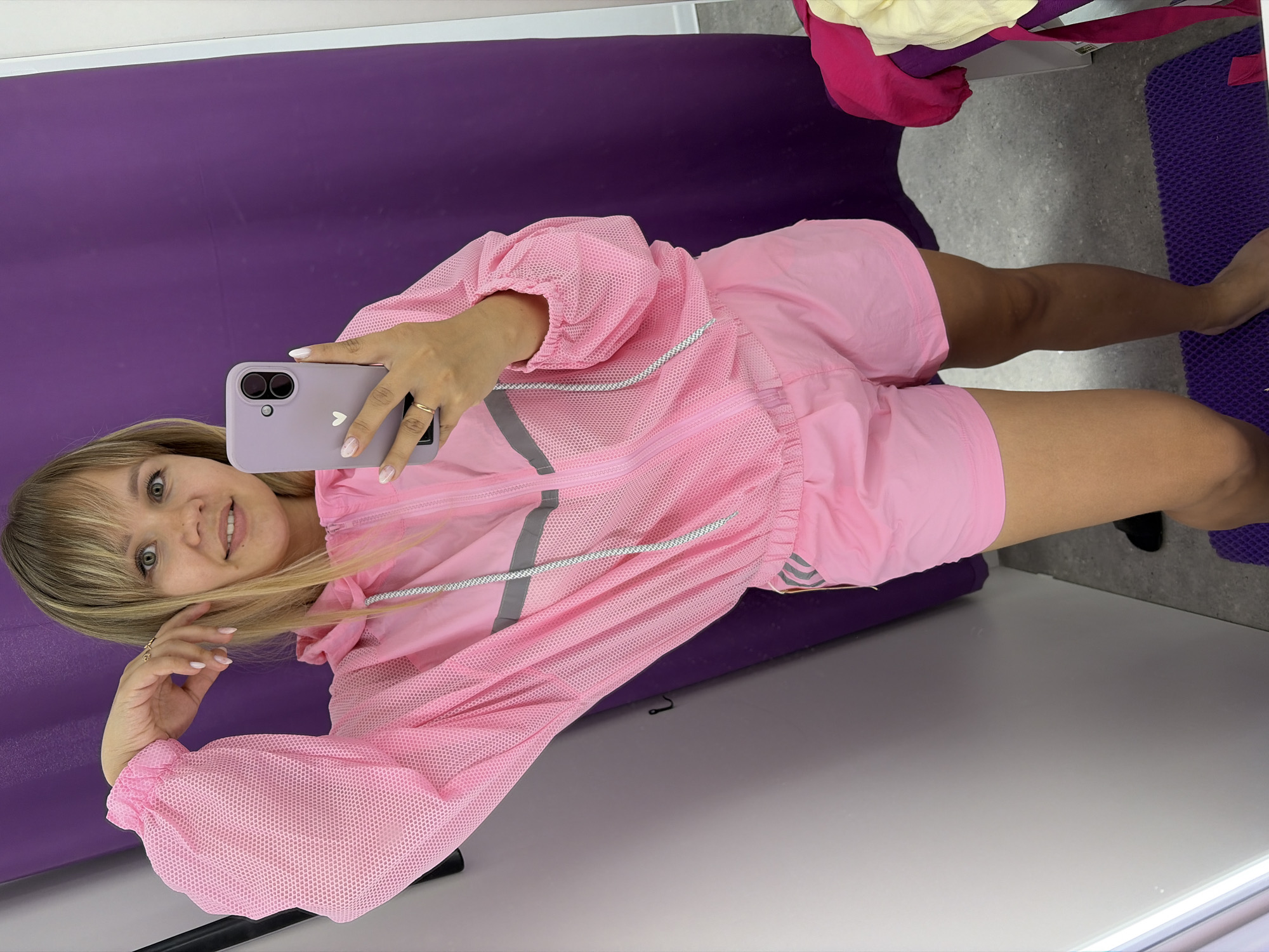 A woman is taking a selfie in a fitting room. She is wearing a pink hooded jacket with a mesh overlay and pink shorts. The jacket has reflective stripes on the chest and sleeves. She is holding a purple smartphone in her right hand, and her left arm is bent at the elbow, resting on her head. She has blonde hair with bangs and is smiling at the camera. The fitting room has a purple curtain in the background and a white door on the left. A gray floor is visible, with a purple mat on the right side of the image. The image appears to be taken in a retail store. There is no information in the image to indicate it is located in Chaikovsky, Russia.