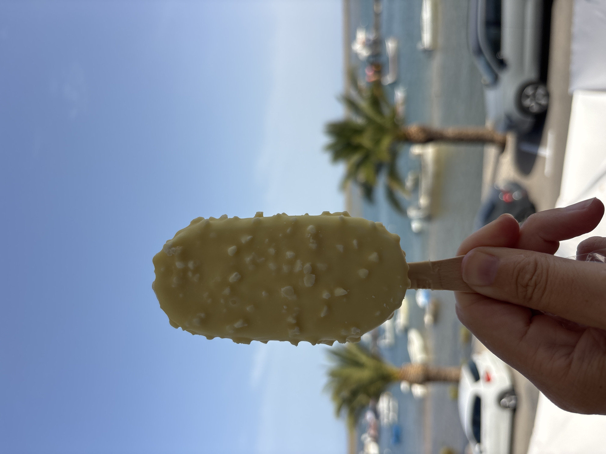 A hand holds a white chocolate-covered ice cream bar with chopped nuts against a bright blue sky. In the blurred background, a body of water, likely the Gilão River in Tavira, Portugal, is visible with several boats. Two palm trees are present on the bank, with cars parked behind them. One car is white, and another is silver.